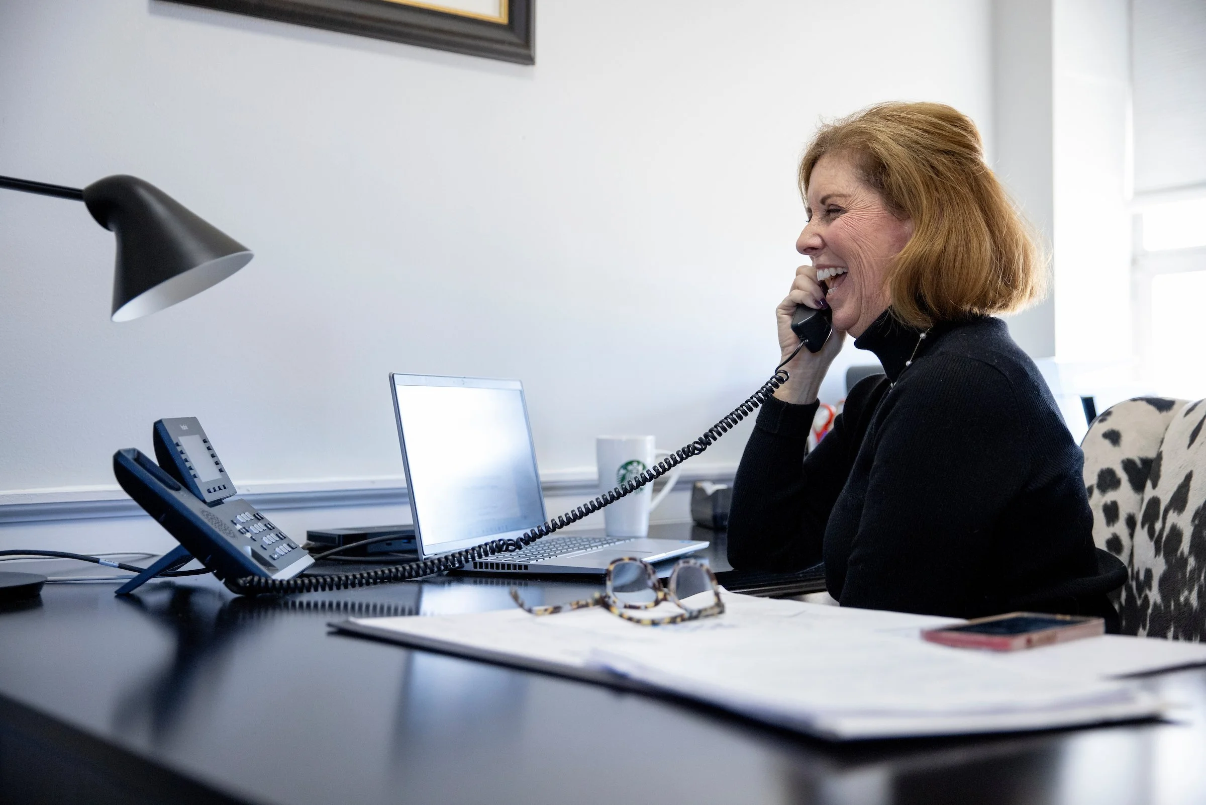 A woman with red hair smiling and talking on a landline phone in an office. The desk has a laptop, a white mug, a pair of glasses, and a calendar, with a black desk lamp and phone on the desk.