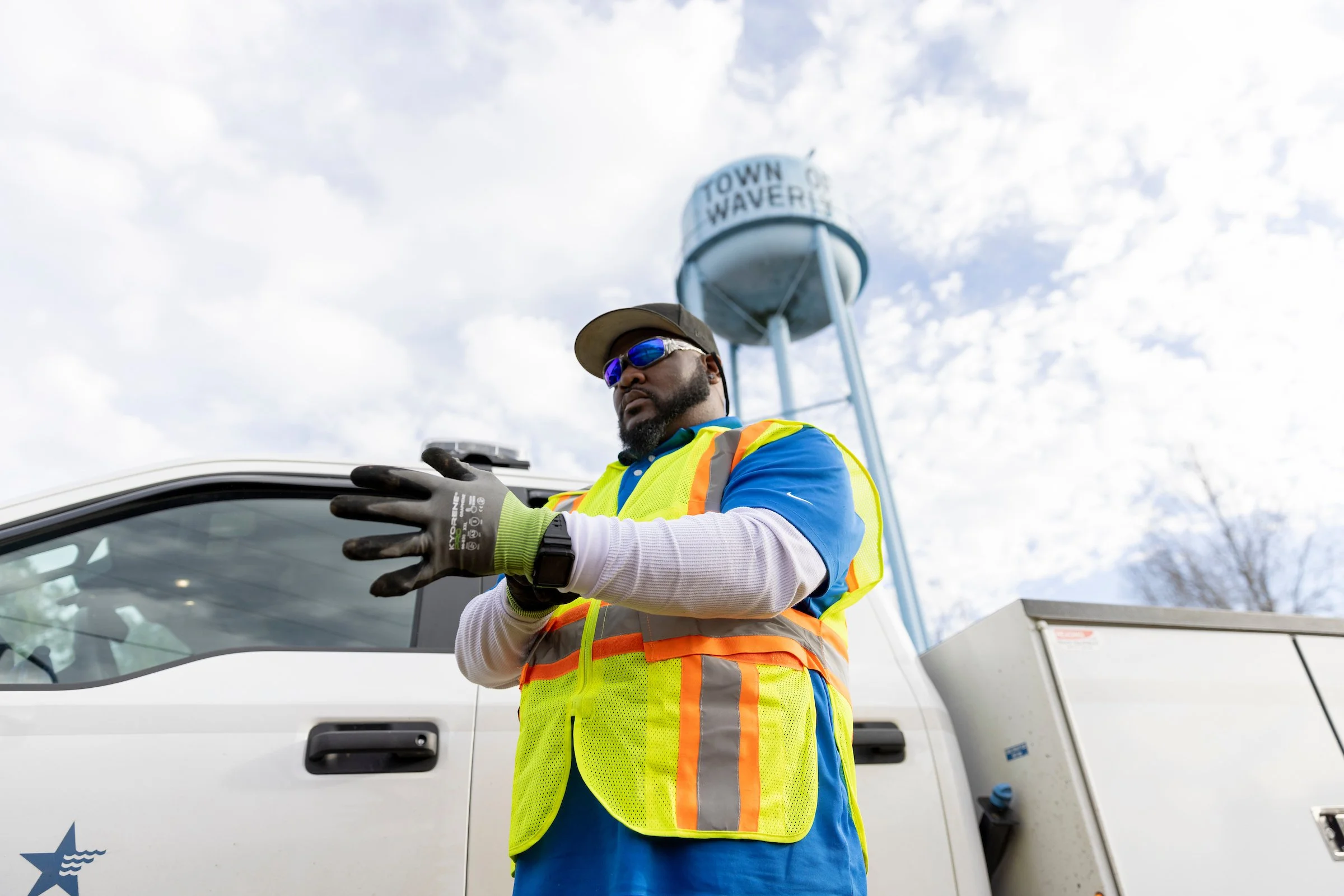 A man wearing sunglasses, a tan hat, a yellow safety vest, and gloves standing in front of a white vehicle with a water tower in the background that has the words "Town of Waverly" on it.