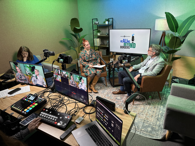 A TV show recording setup with three people seated in a studio, two women and one man, surrounded by cameras, monitors, and electronic equipment, with a large screen displaying the show title 'Healing Beyond Borders' and decorative plants in the back