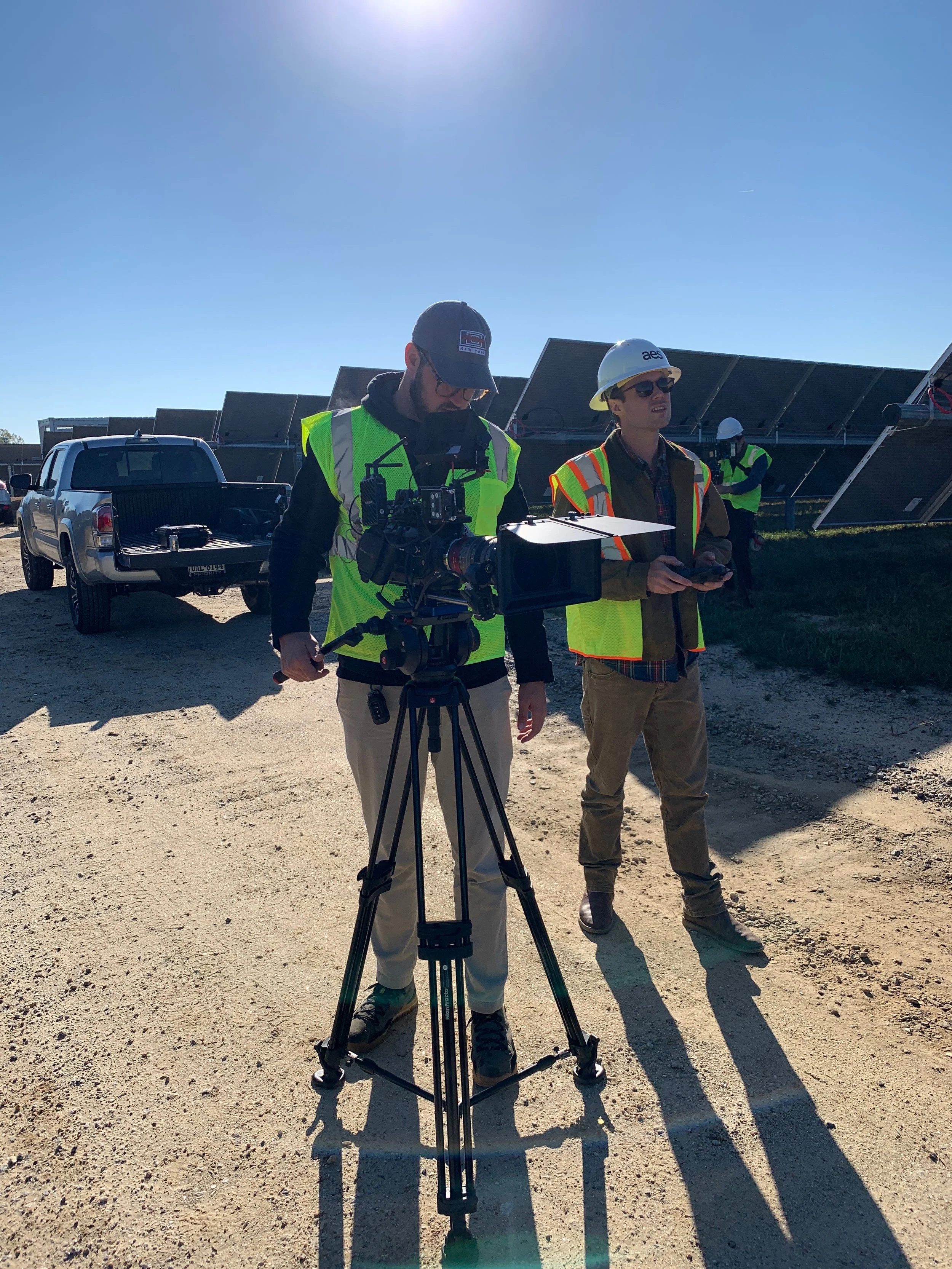 Two men wearing safety vests and hard hats working on solar panel installation or maintenance outdoors, with solar panels in the background and vehicles nearby.
