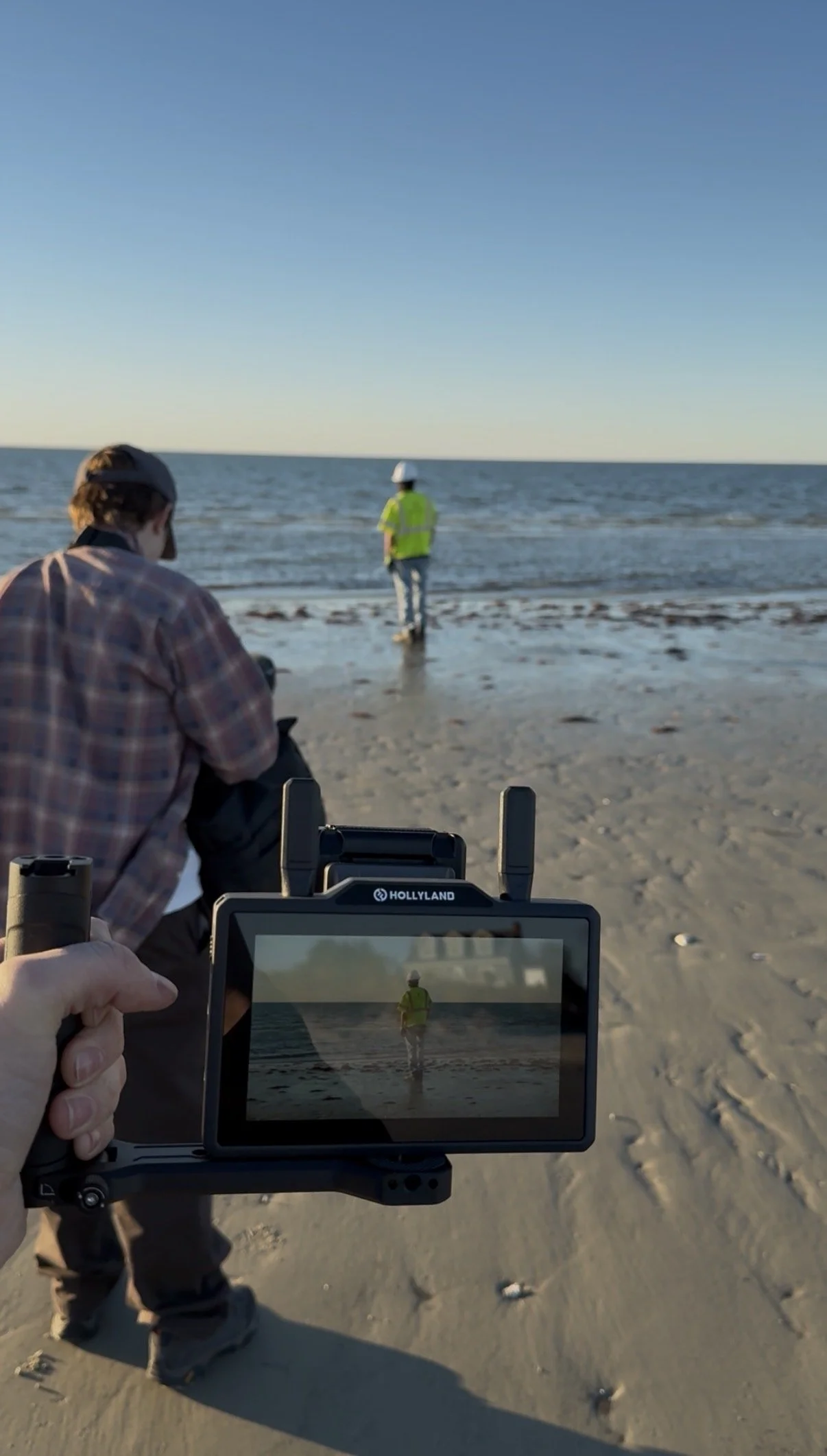 Photograph of a person holding a remote control, filming a man in a yellow safety vest and white hard hat standing near the shoreline at the beach, with the ocean and clear blue sky in the background.