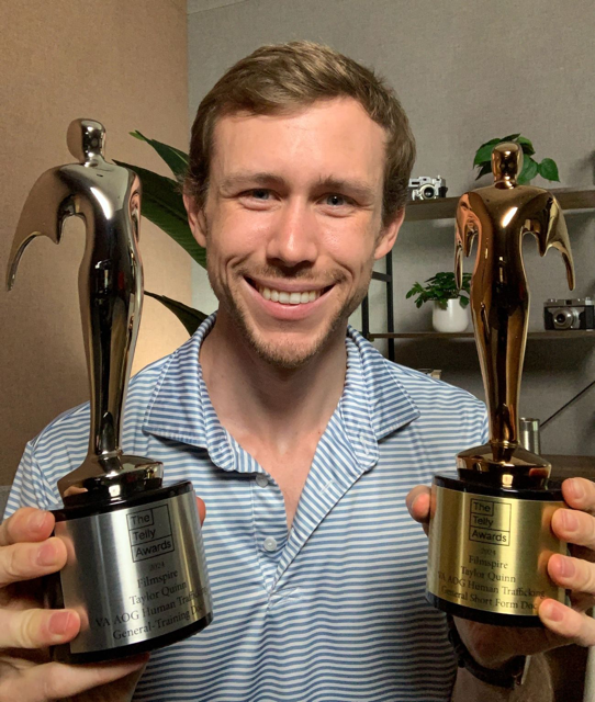 A man smiling and holding two awards with a figurine on top, in an indoor setting with plants and shelves in the background.
