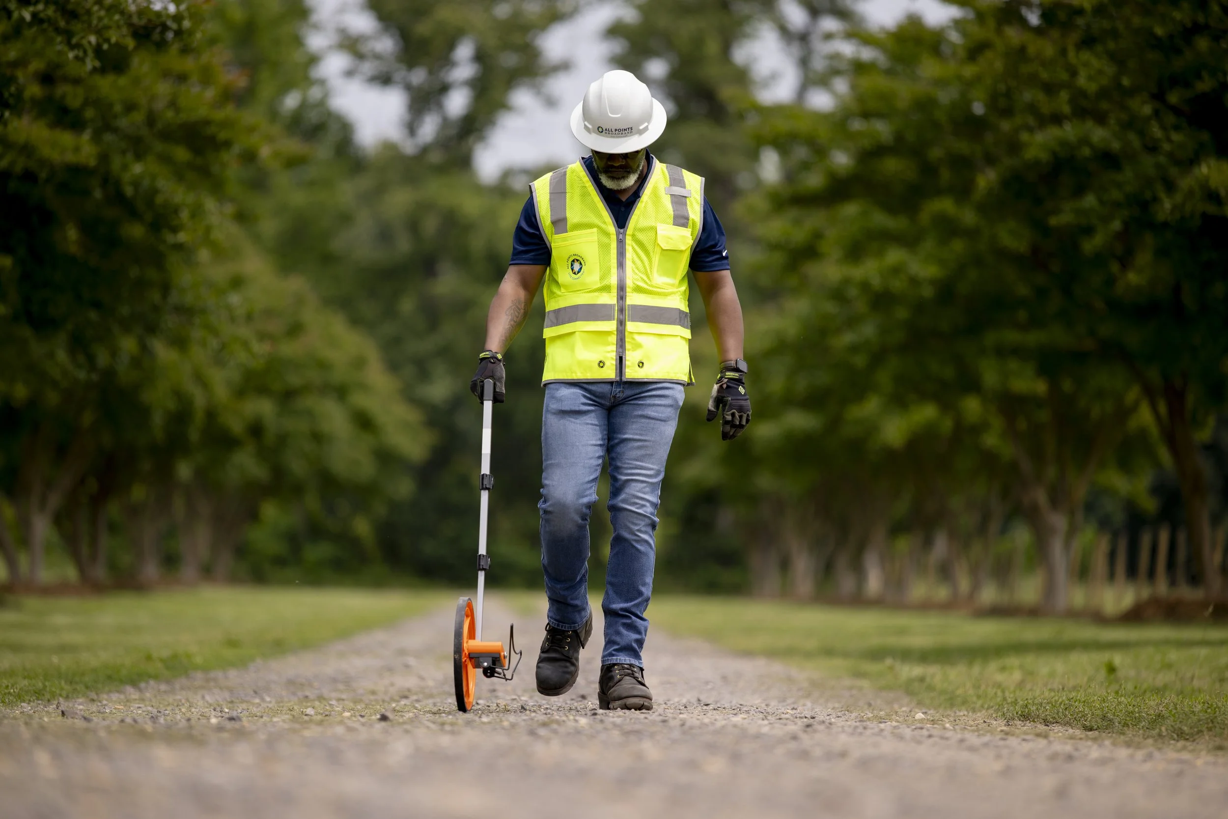 A man in a yellow safety vest, white helmet, and gloves is walking on a gravel path with a metal detector. He is outdoors, surrounded by green trees.
