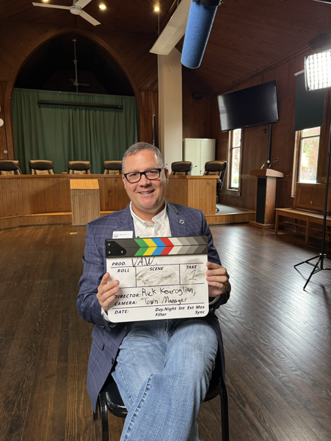 Man in a suit holding a film slate inside a wooden hall with a stage and green curtains.
