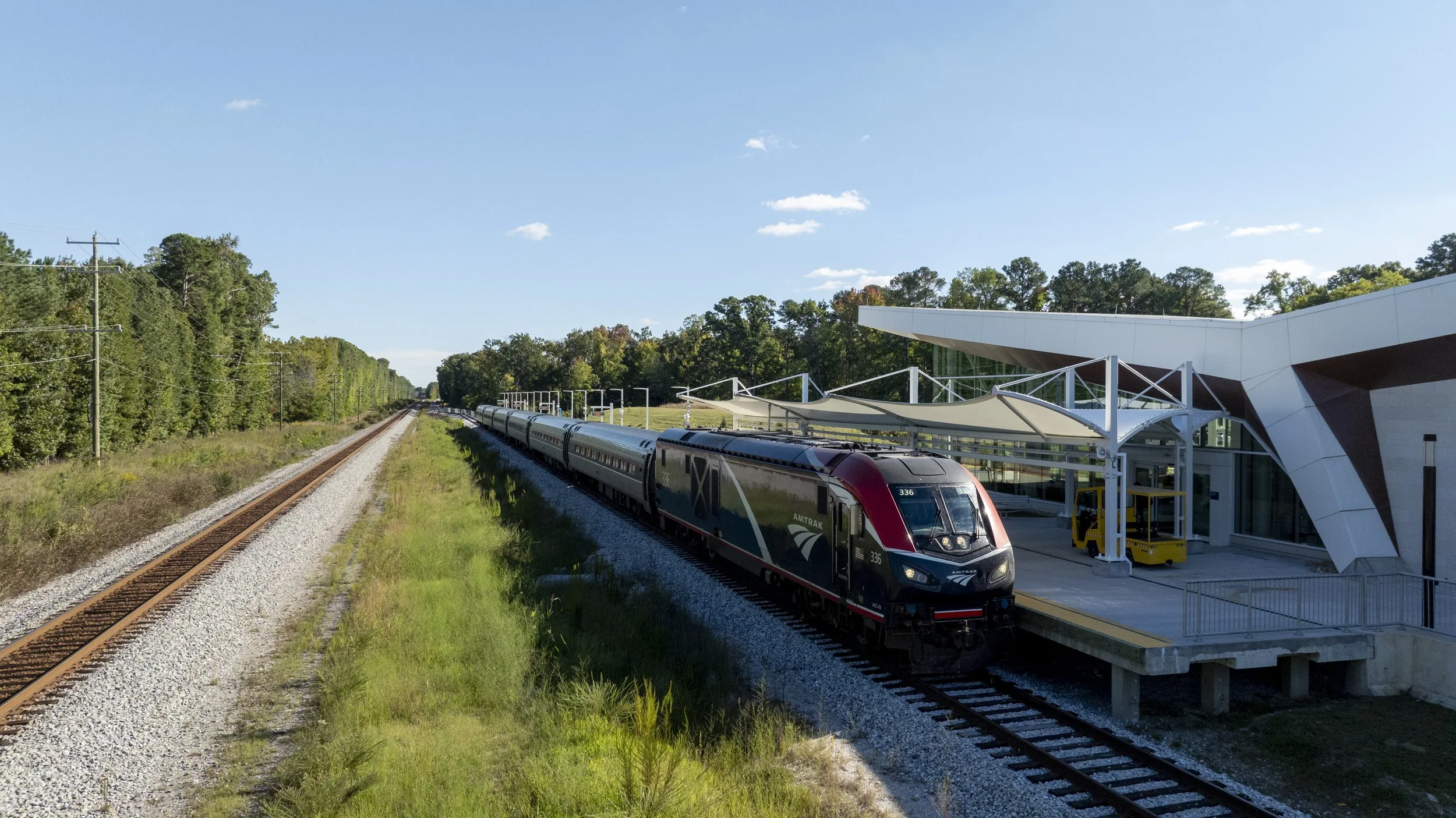 A modern train at a station next to a wooded area on a sunny day.
