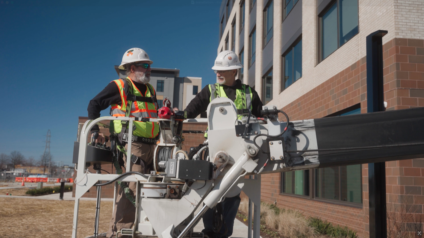 Two construction workers with safety helmets and vests working on a boom lift near a multi-story brick building under a clear blue sky.