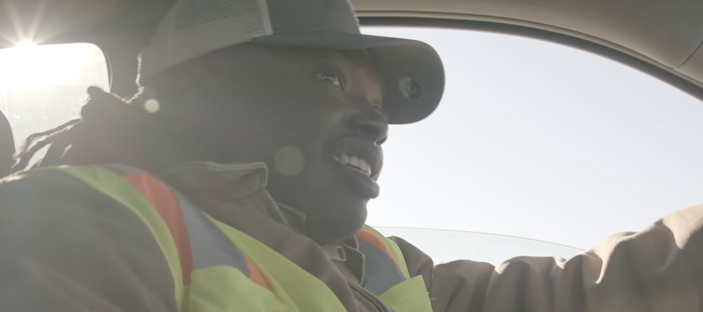 A smiling man wearing a high-visibility safety vest, a jacket, and a baseball cap, possibly a construction or road worker, sitting inside a vehicle with sunlight shining in.