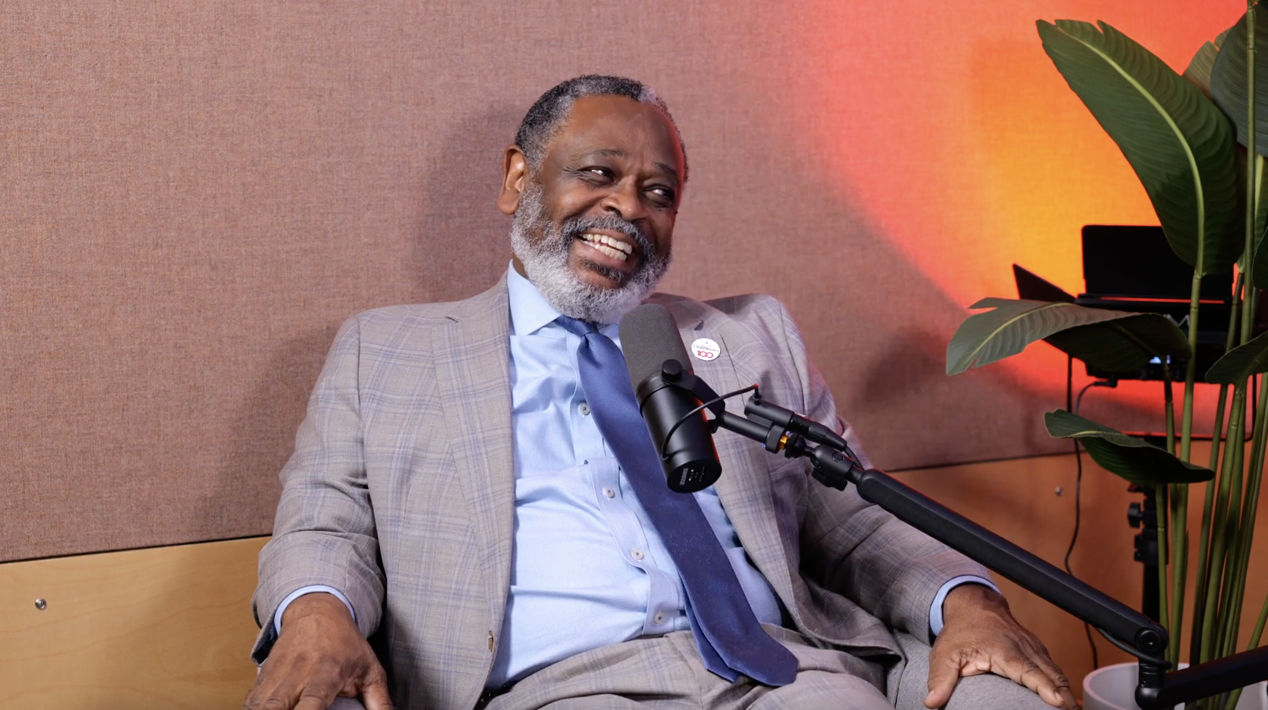 An elderly man with gray hair and a beard, wearing a light gray plaid suit and blue tie, sitting in a room with beige walls and a large green plant, smiling and speaking into a microphone during a podcast or interview.