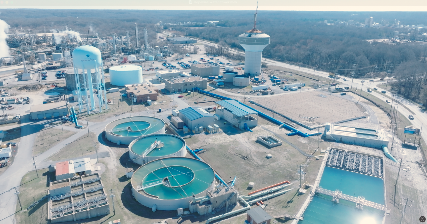 Aerial view of a water treatment plant with circular clarifiers, large storage tanks, and a tall control tower, surrounded by roads and a wooded area.