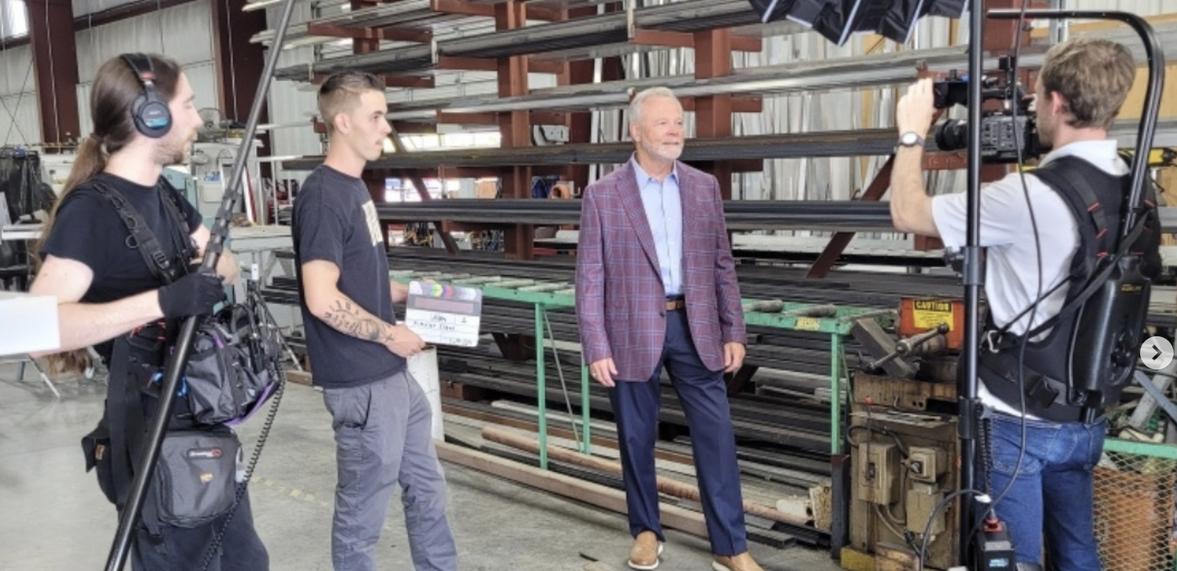 A film crew is recording an interview or scene with an older man in a warehouse or industrial setting. The crew members are holding filming equipment, and the older man is standing near metal pipes or beams, smiling.