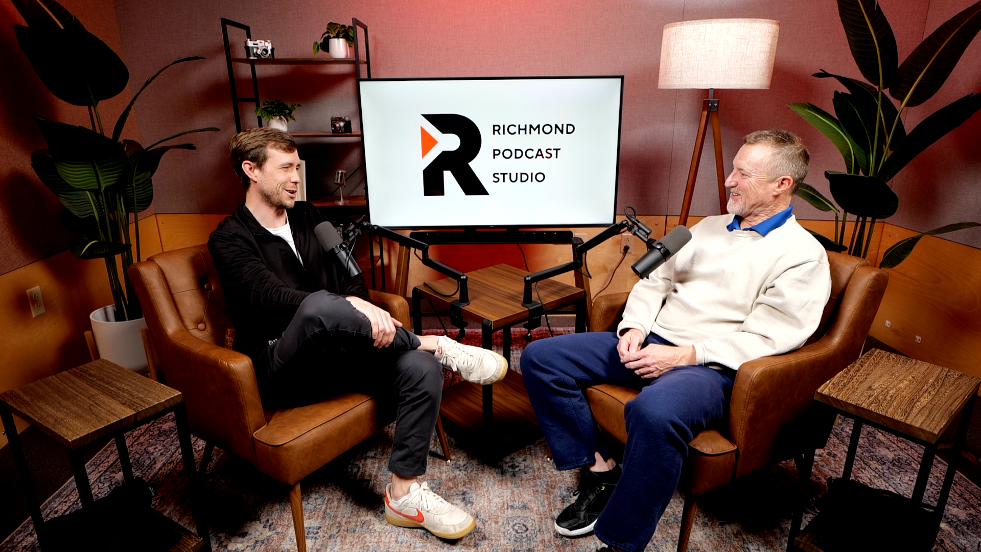 Two men sitting on leather chairs in a podcast recording studio, speaking into microphones with a large screen behind them displaying the text 'Richmond Podcast Studio' and a logo.