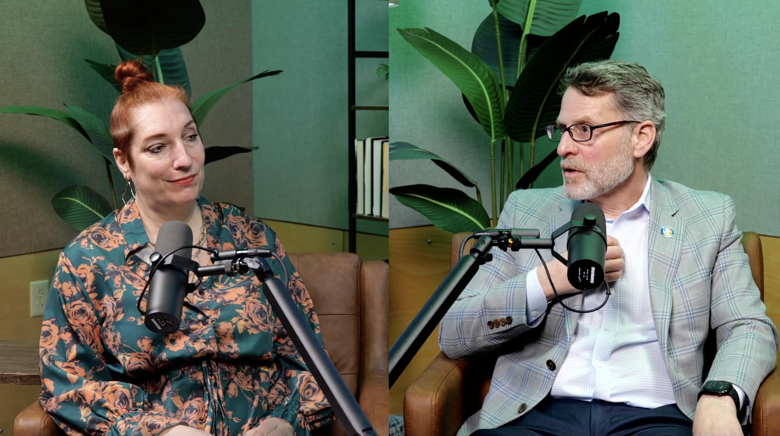 Two people sitting at a table with microphones, engaged in a conversation. The woman on the left has red hair in a bun, wearing a floral blouse. The man on the right has gray hair and beard, wearing glasses and a light checkered blazer.