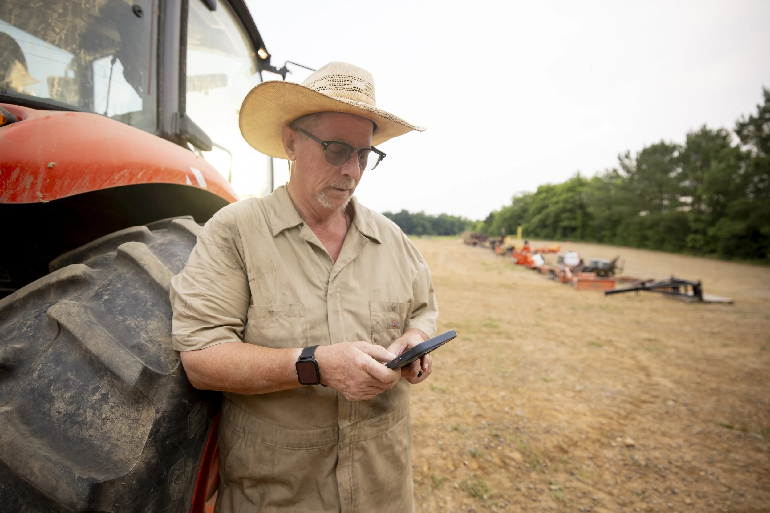 A man with glasses, a beige shirt, and a straw hat standing next to a large tractor, looking at a smartphone in his hands in an open field with trees and farming equipment in the background.