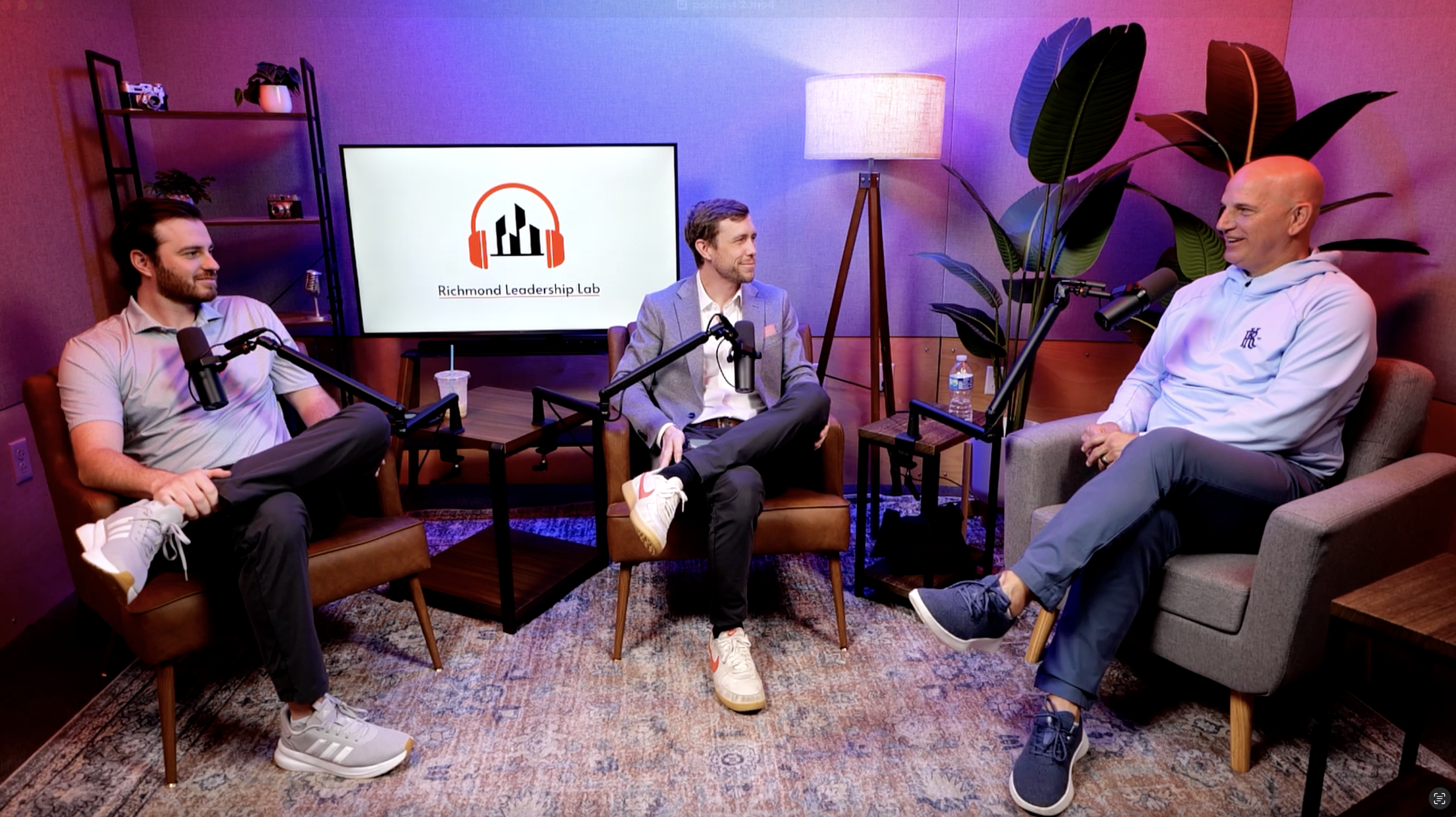 Three men sitting in a podcast recording studio having a conversation, with microphones in front of them. A large screen in the background displays the text 'Richmond Leadership Lab' with a logo of headphones and city skyline. The studio has warm lig