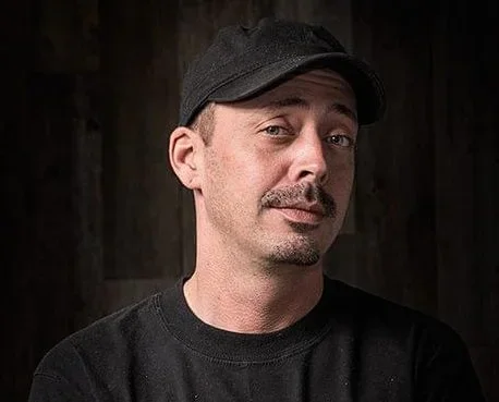 A man wearing a black cap and black shirt against a dark background, looking at the camera with a slight smirk.