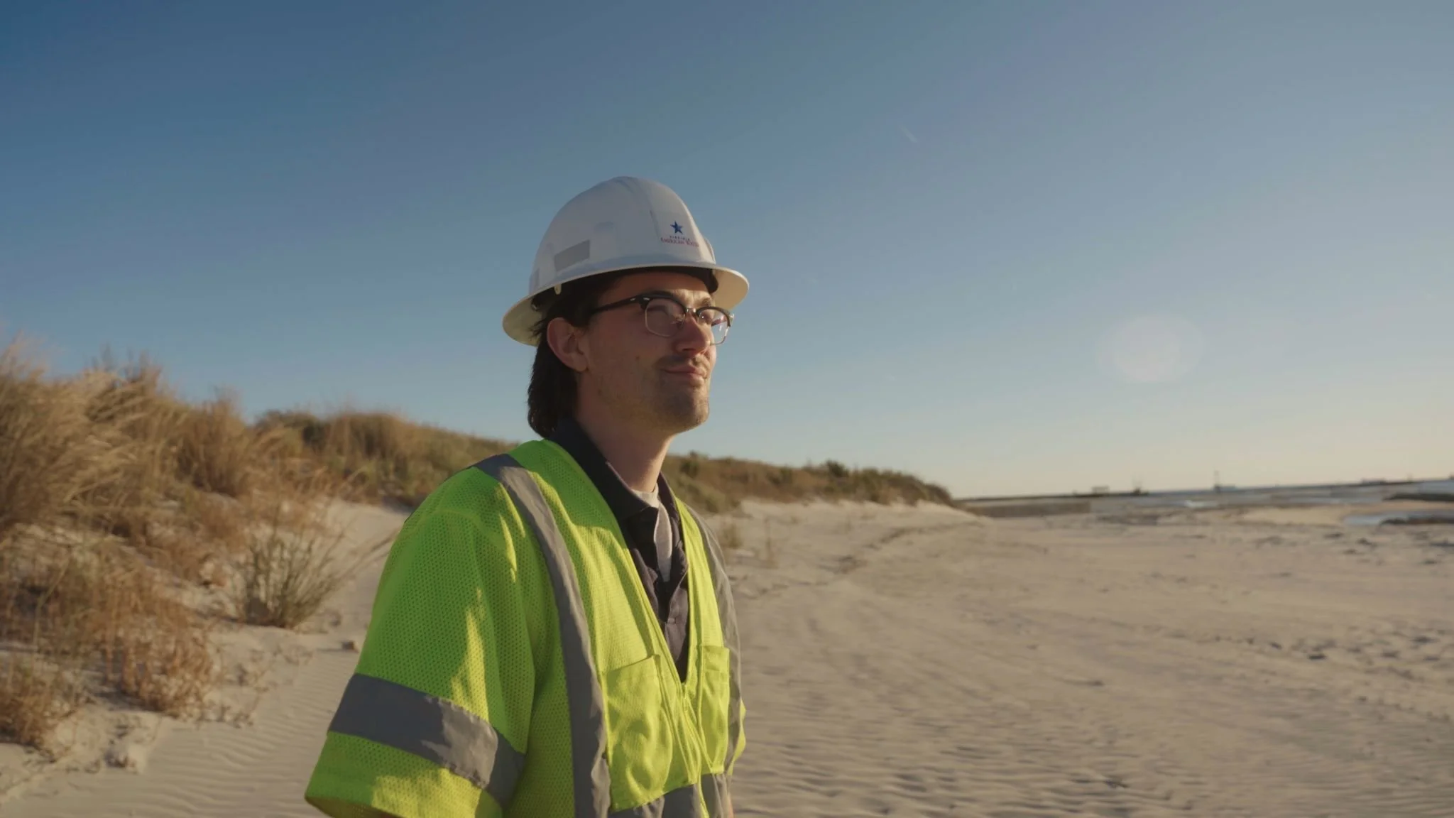Man wearing a white safety helmet and yellow reflective vest walks on a sandy beach with dunes and sparse vegetation under a clear blue sky.