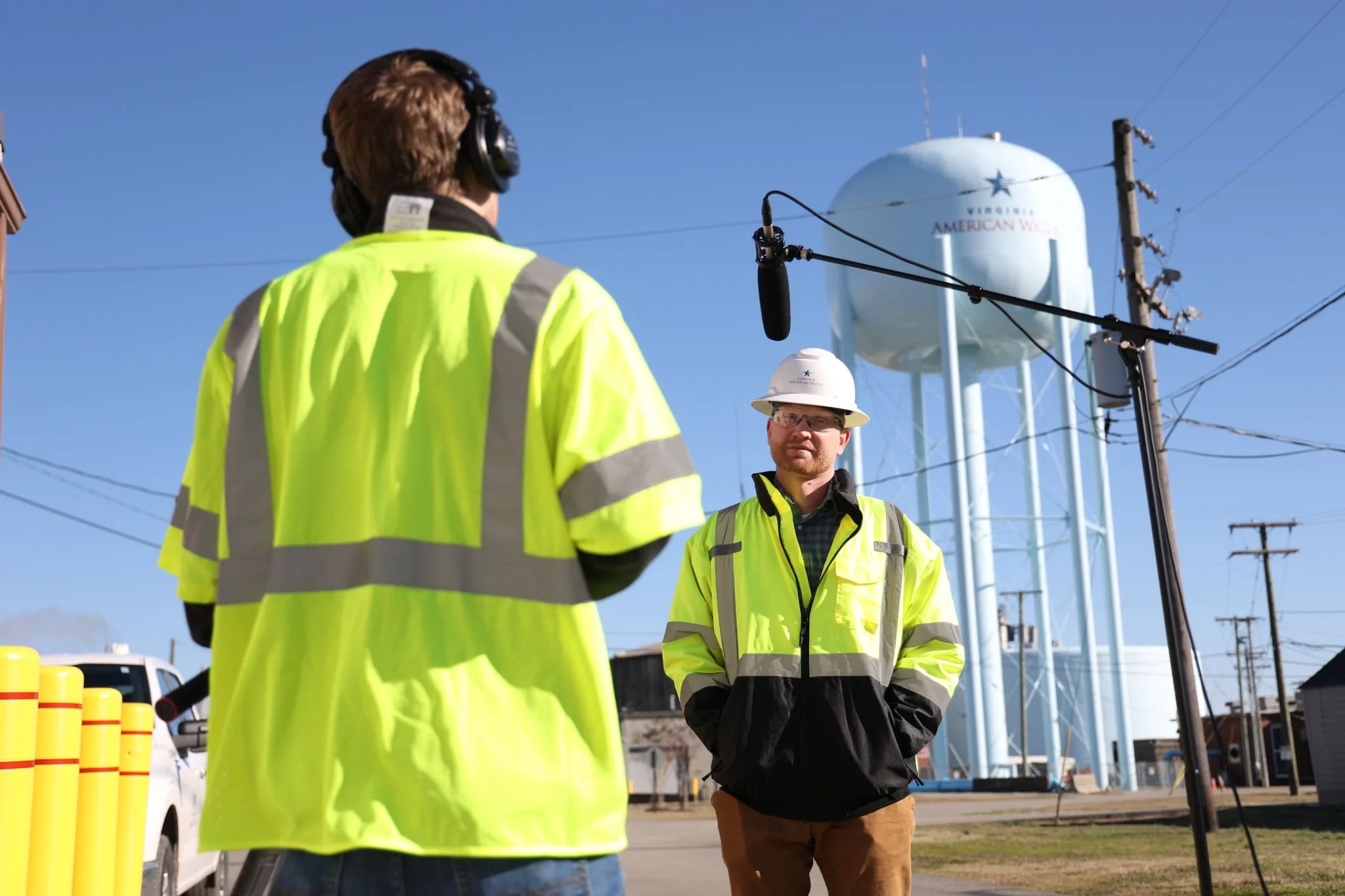 Two construction workers in high-visibility jackets and hard hats stand outdoors near power lines and utility poles, with a water tower in the background.