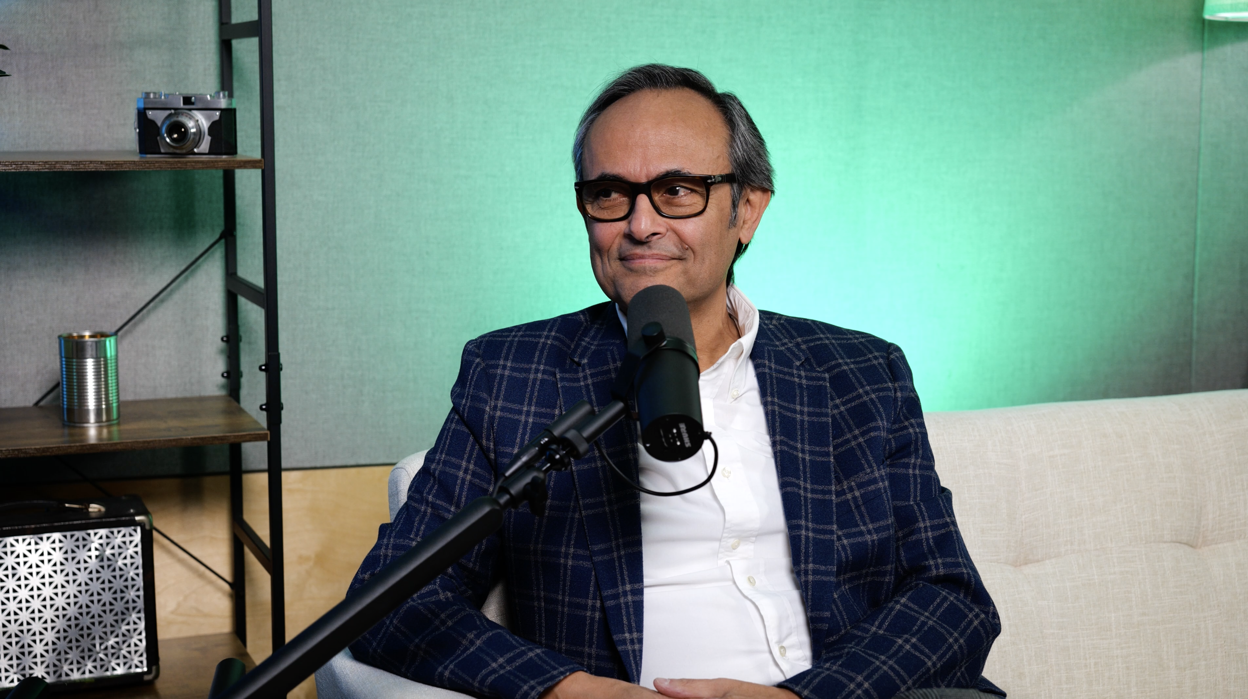A man with gray hair and glasses sitting in front of a microphone in a studio setting, wearing a checkered blazer and white shirt, with a green background and shelves with a camera and cans behind him.