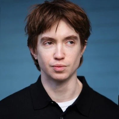 A young man with brown hair wearing a black shirt against a blue background.