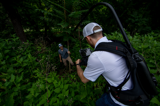 Photographer taking pictures of a man in a forested area surrounded by green foliage.