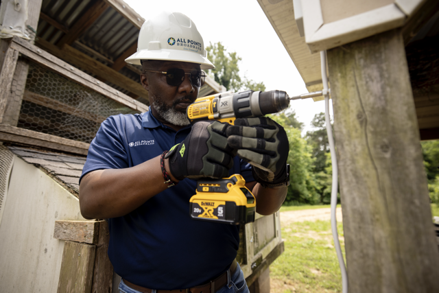 A man wearing sunglasses, a white safety helmet, and a blue shirt labeled 'All Points Broadband' is using a cordless drill to work on an outdoor wooden structure.