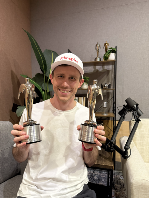 Man smiling, wearing a white cap with 'Filmspire' logo, holding two gold statue awards in a living room with a plant and a shelf with trophies and camera in the background, and a microphone stand in the foreground.