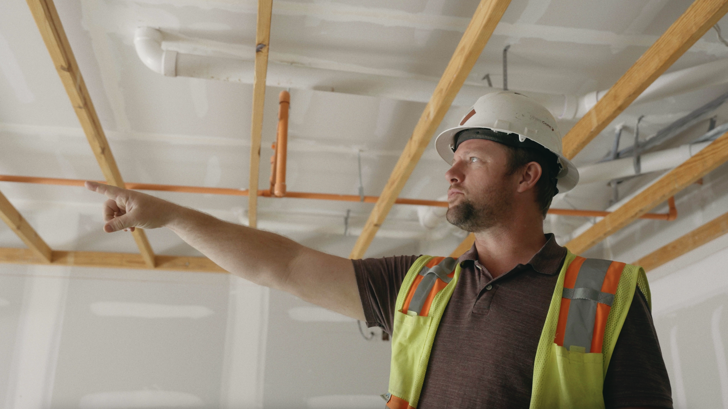 A construction worker in a hard hat and safety vest gesturing and pointing upward at a ceiling under construction, with exposed wooden beams and pipes.