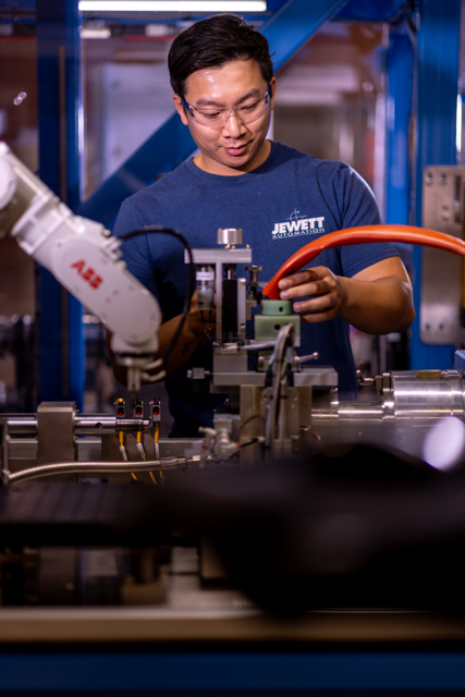 A technician working with machinery in a lab or manufacturing environment, wearing safety glasses and a blue shirt with Jewett Automation printed on it.