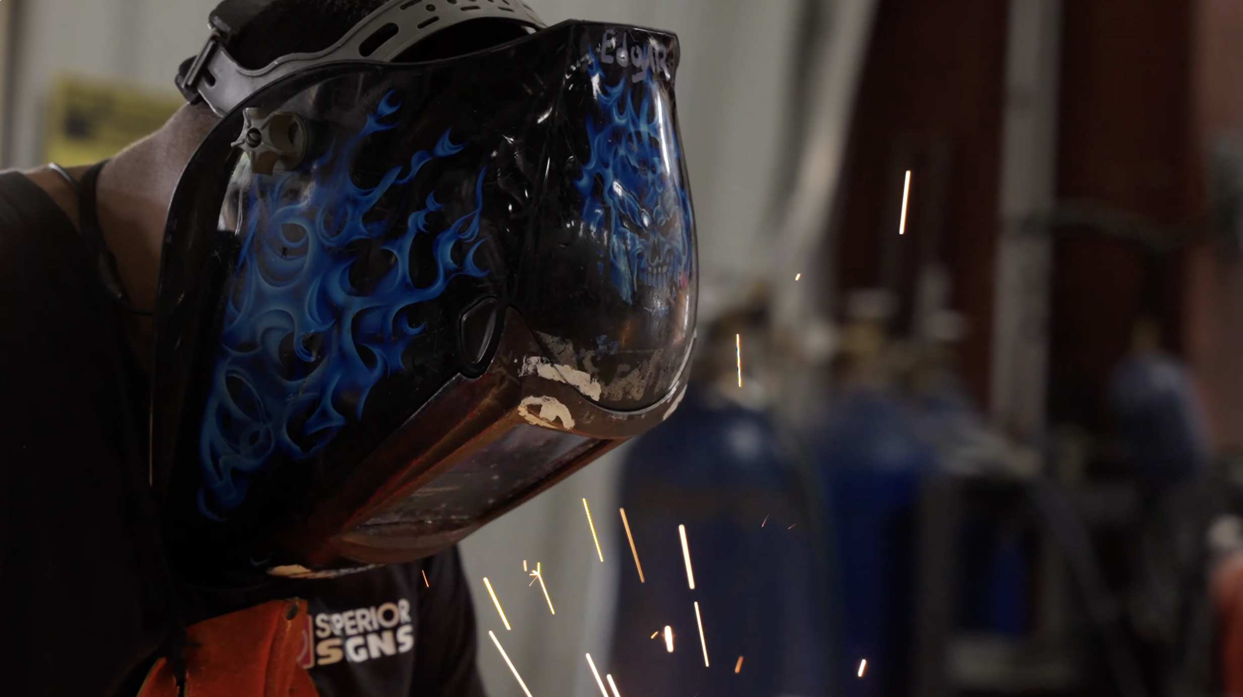 A person welding with a helmet featuring blue flame design, sparks flying from the welding process, and a blurred workshop background.