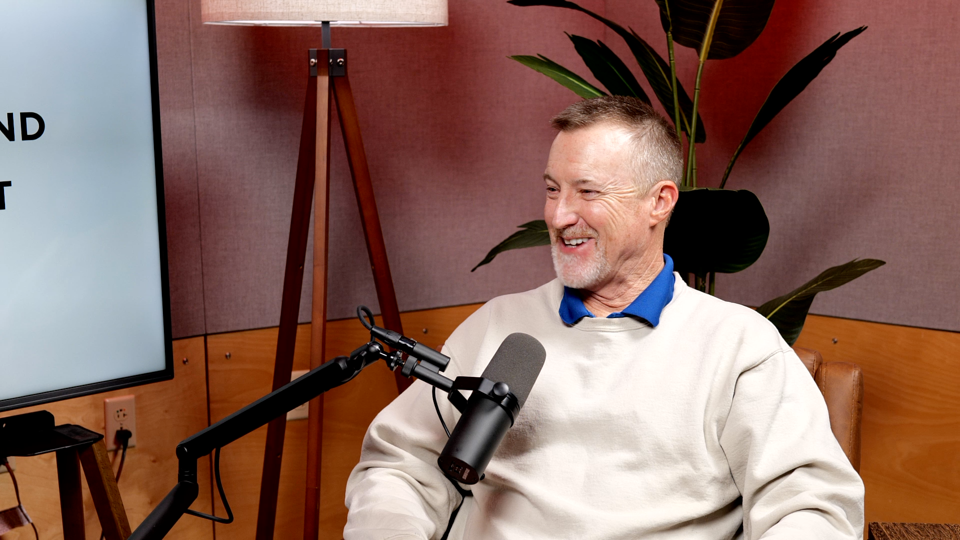Smiling man with short hair and beard, wearing a white sweater over a blue collared shirt, sitting at a podcast microphone in a room with a large screen, wooden walls, a floor lamp, and large green plant.
