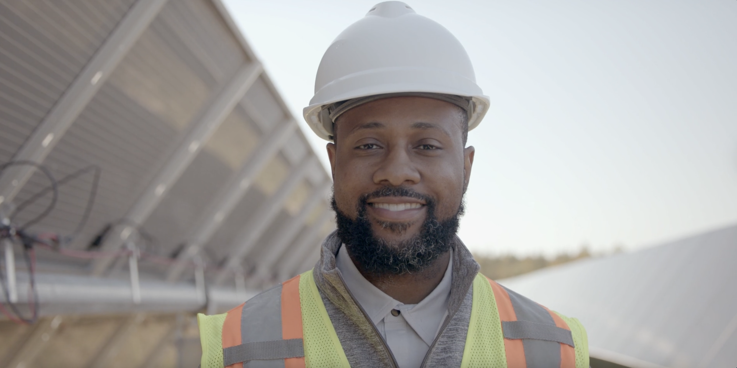 Man wearing a white safety helmet and a neon orange safety vest smiling at the camera, with solar panels in the background.