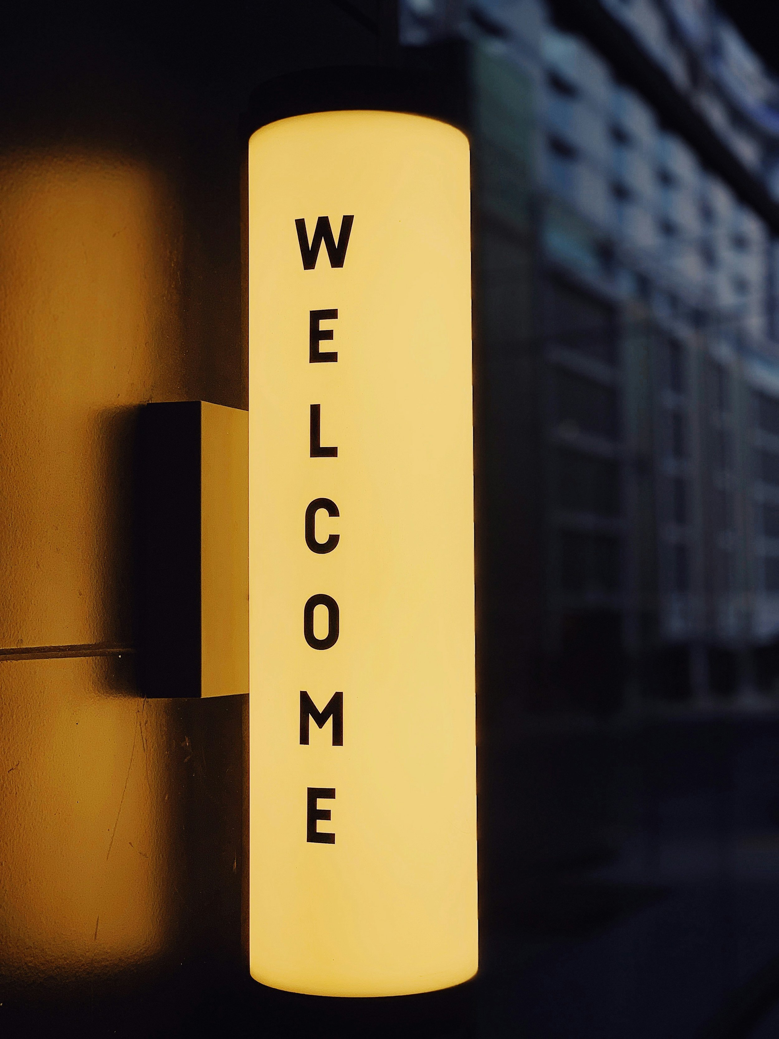 A tall, cylindrical, illuminated sign with yellow background and black letters spelling 'WELCOME' vertically. The sign is mounted on a wall next to an air conditioning unit.