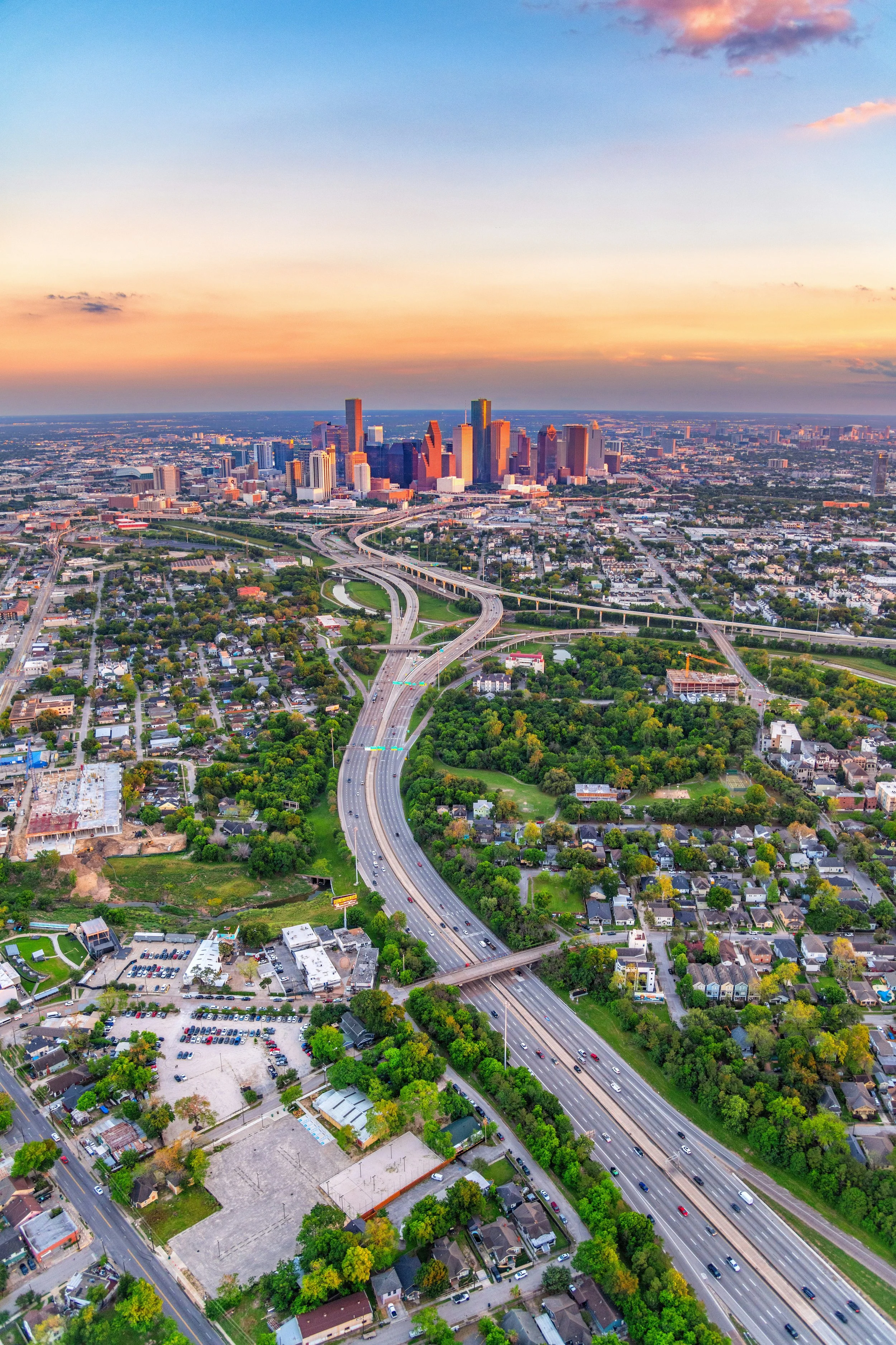 An aerial view of a city skyline at sunset, with skyscrapers, highways, and residential areas with trees and houses.