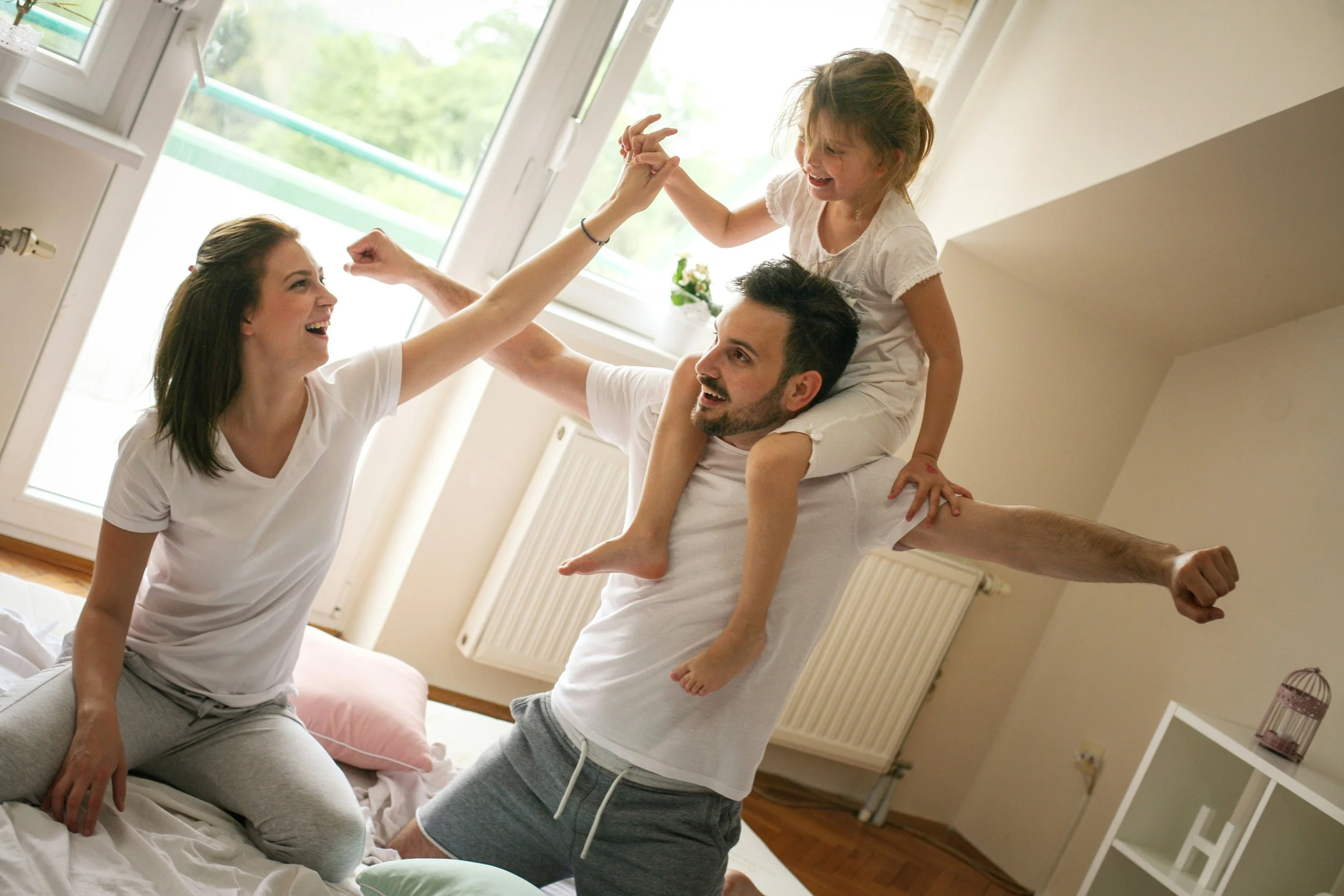 A family having fun together in a bedroom, with a young girl sitting on her father's shoulders and holding his hand, while her mother sits on the bed smiling and raising her fist in celebration.