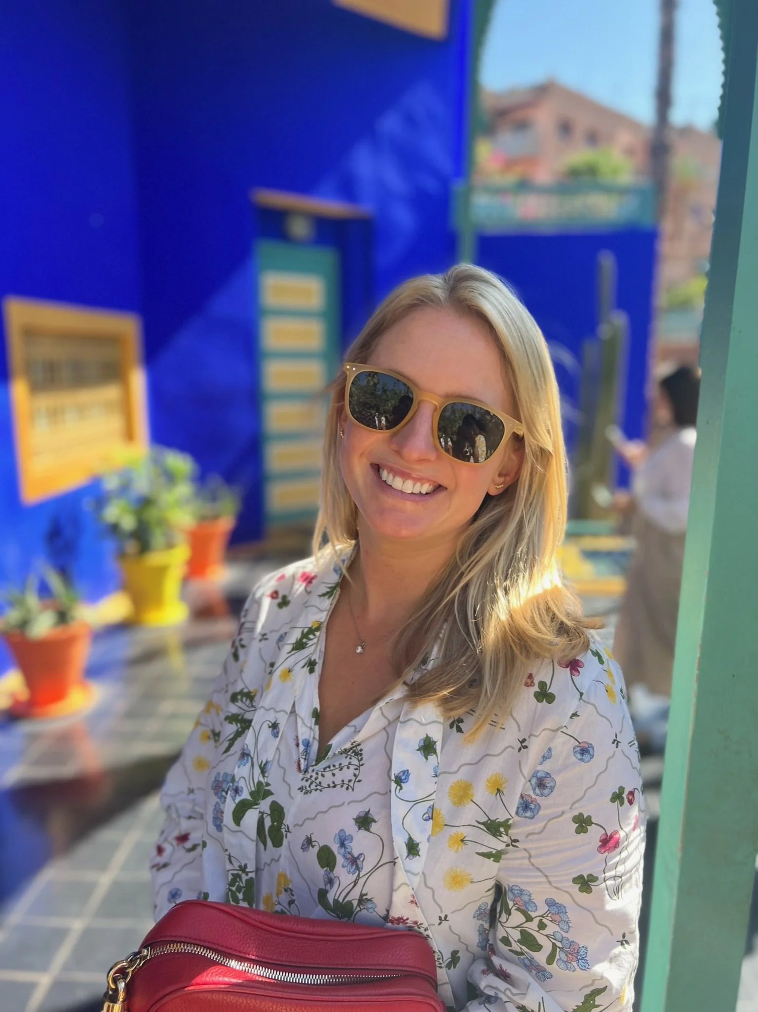 A woman with blonde hair wearing sunglasses and a floral blouse smiling outdoors, with colorful pots and plants in the background.