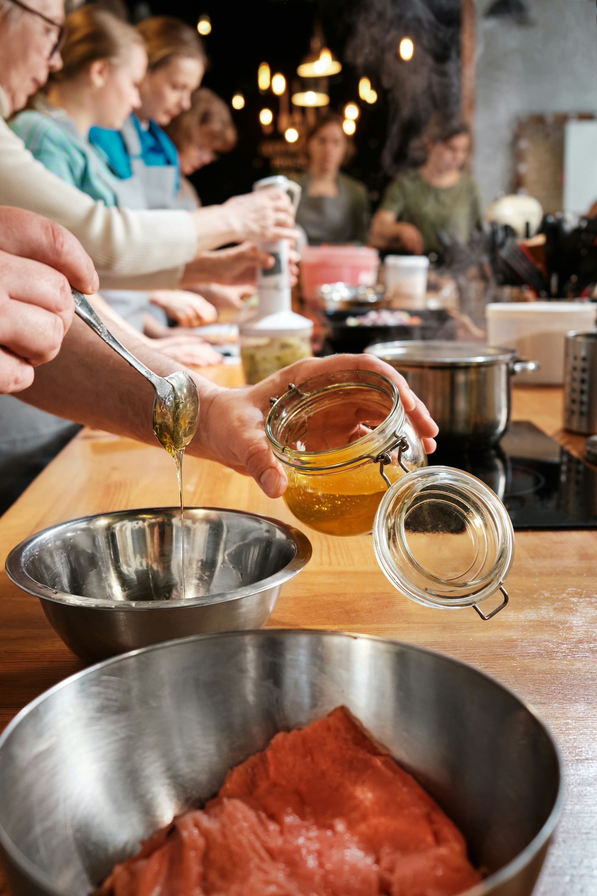 People in an indoor kitchen preparing ingredients for cooking, with various food items, jars, and kitchen utensils on the wooden counter.