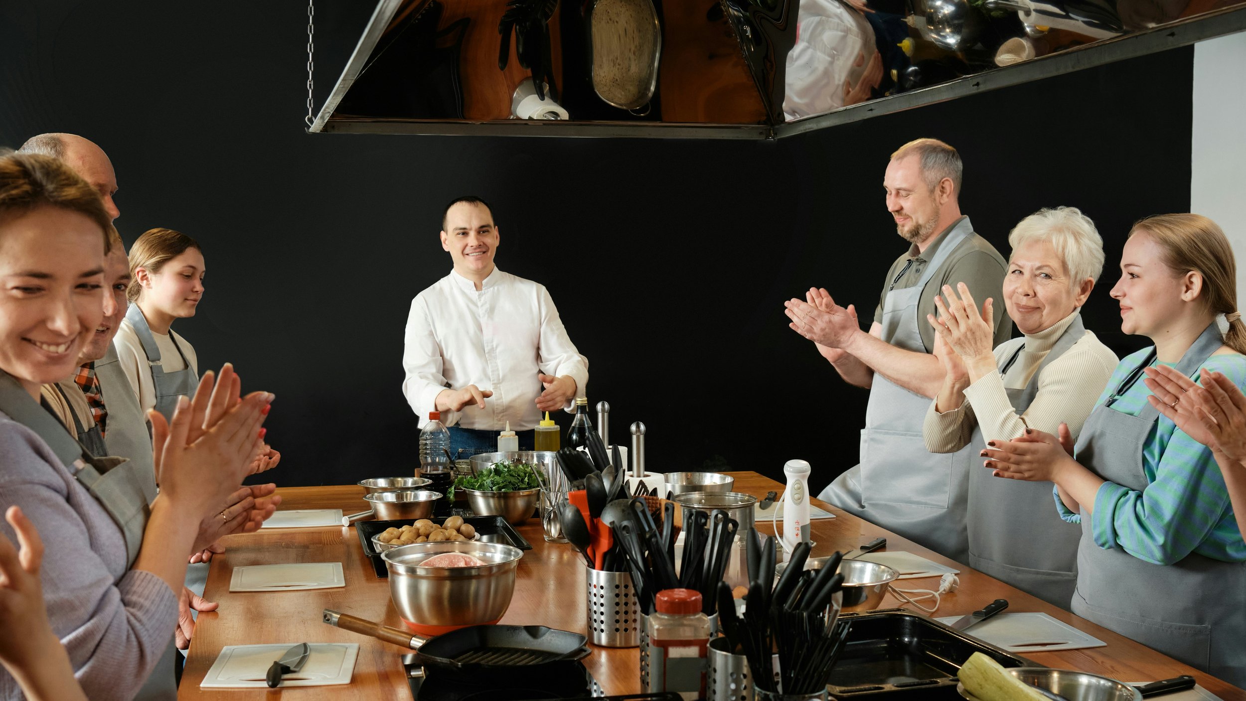 Group of people in aprons standing around a kitchen counter, clapping and smiling, during a cooking class or demonstration.