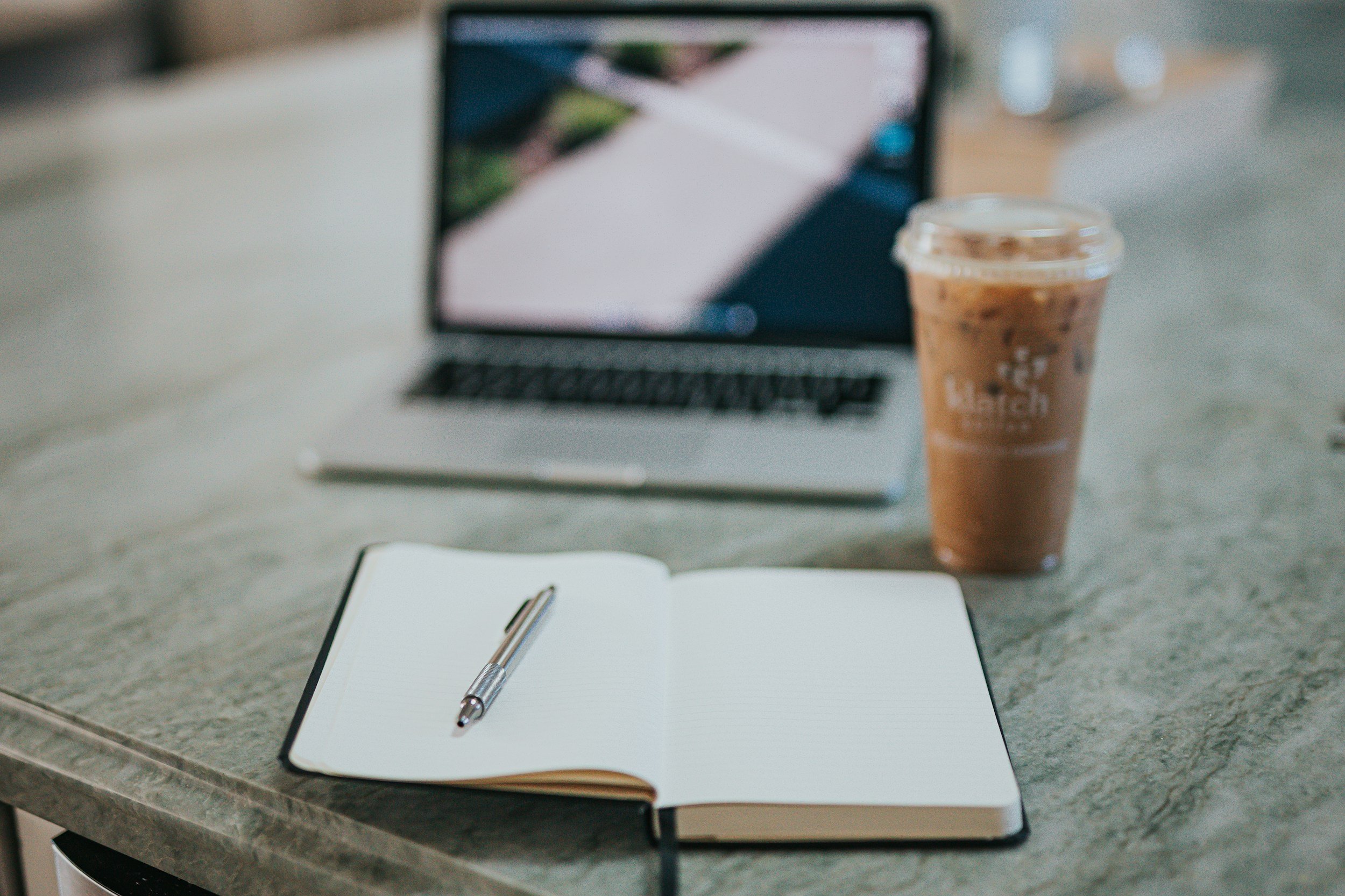 A notepad with a pen, an open book, a laptop, a cup of iced coffee, and a smartphone on a kitchen countertop.