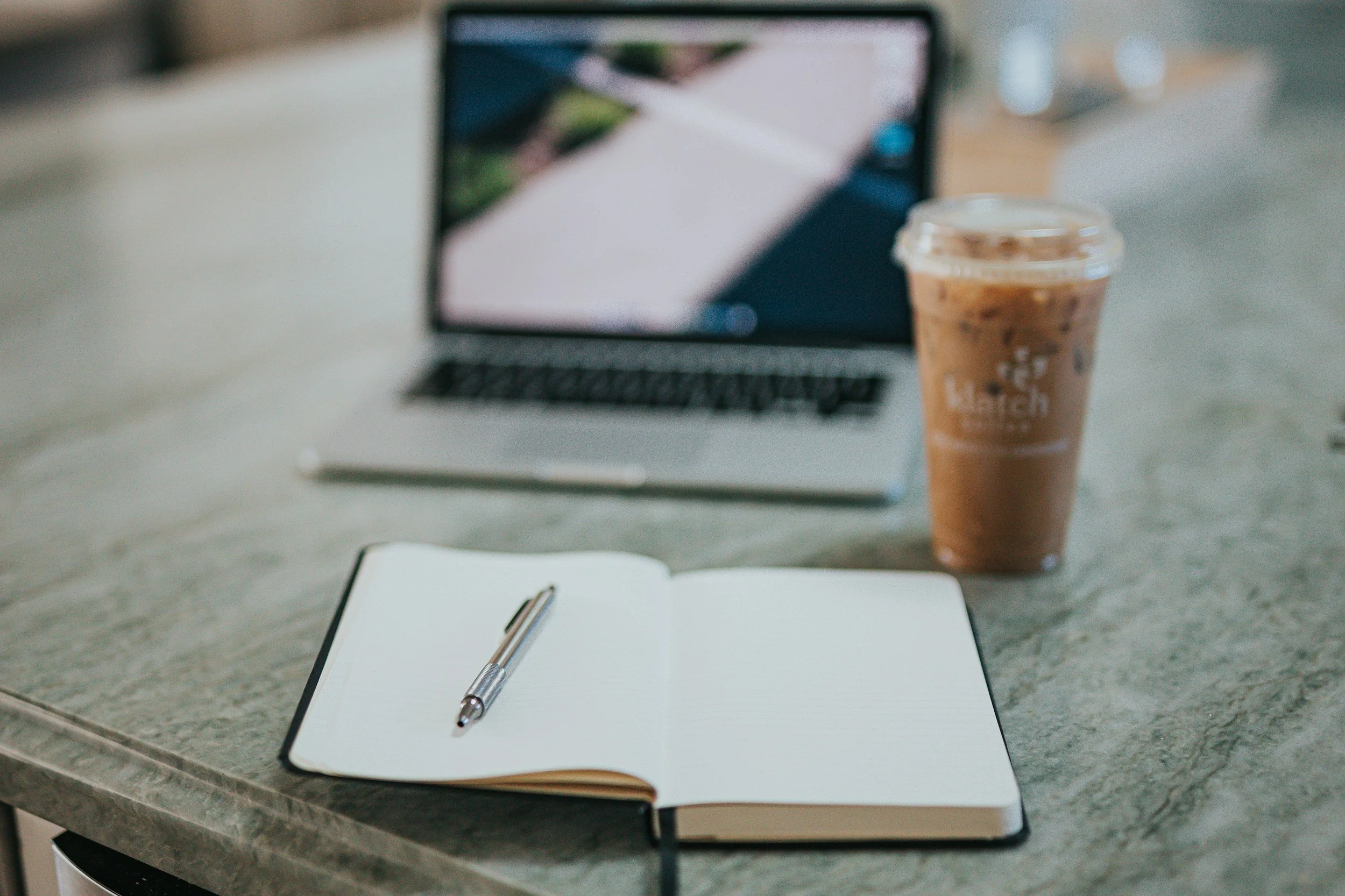 A blurred image of a workspace with a laptop, a coffee cup, an open notebook with a pen, and a smartphone on a gray surface.
