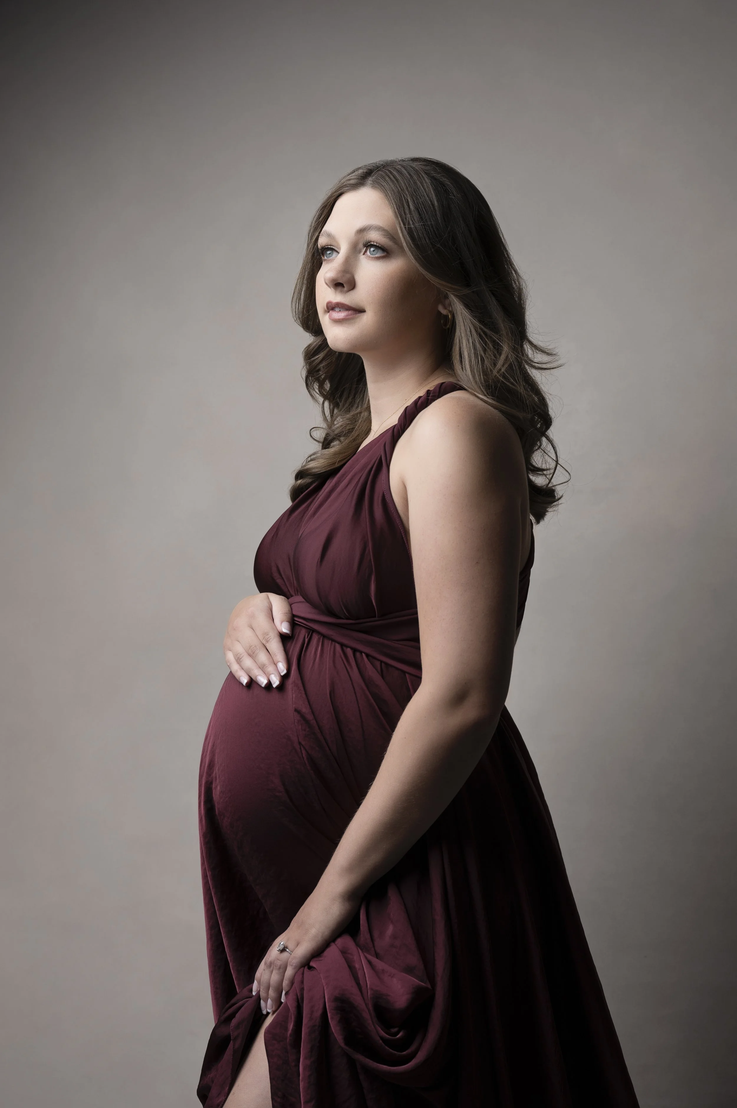 A pregnant woman with long brown hair and blue eyes wearing a strapless, deep burgundy gown with a ruched bodice, standing against a neutral background. She is gently holding her belly with her right hand and looking slightly upward with a peaceful expression.