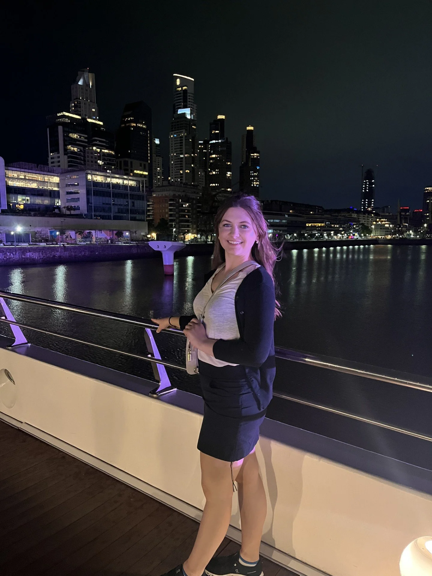 A woman smiling in front of city skyscrapers at night on a boat deck, with illuminated buildings reflected in the water.