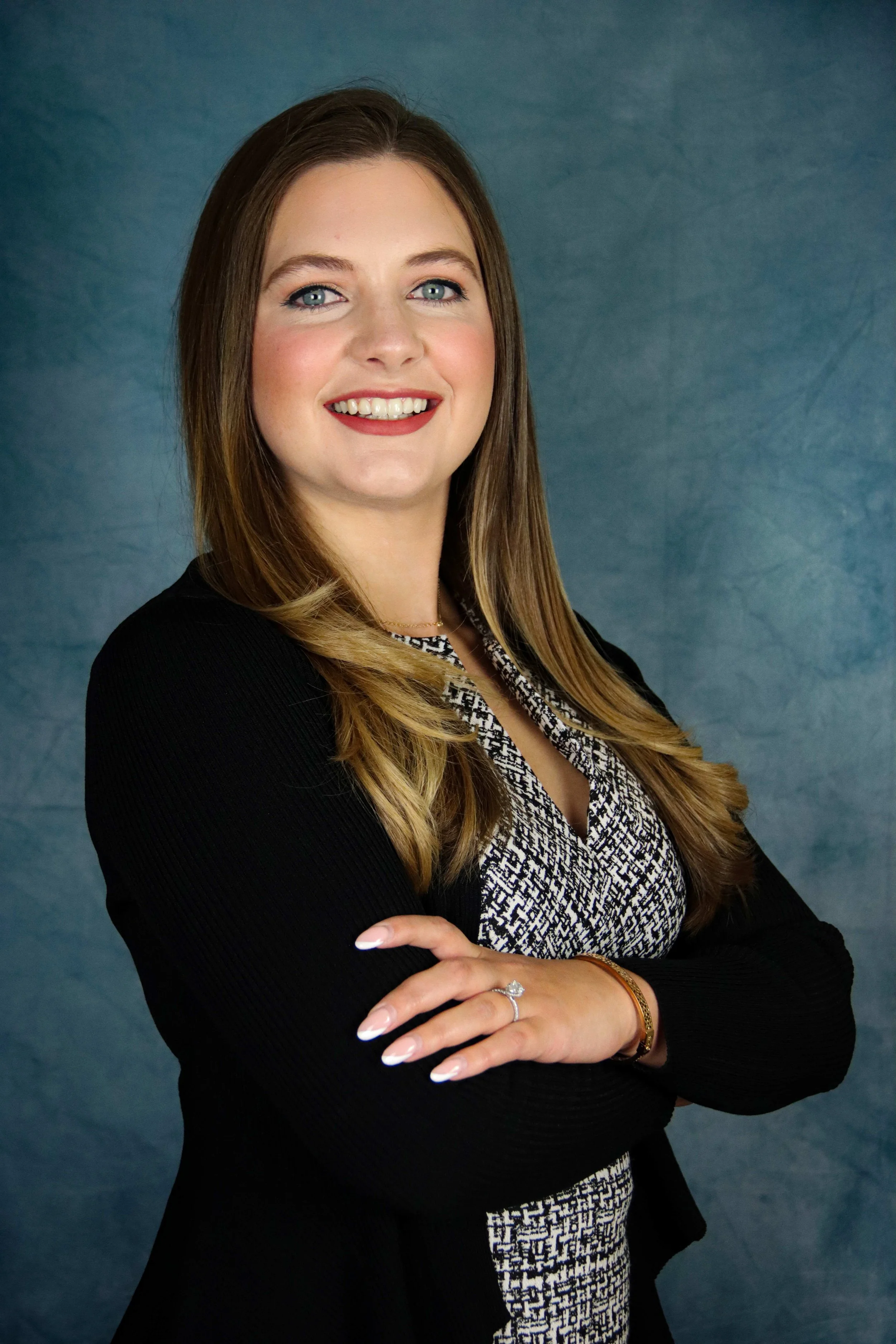 A woman with long, wavy, light brown hair, light blue eyes, and fair skin standing with her arms crossed smile at the camera, wearing a black blazer over a black and white patterned dress, with a blue background.