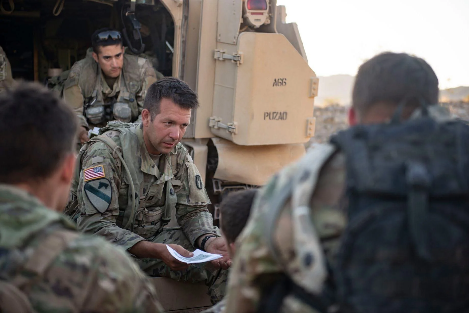 A group of soldiers in military uniforms gathered around a soldier holding a paper, sitting on the ground near a military vehicle, with one soldier sitting inside the vehicle, during what appears to be a briefing or discussion in a desert-like environment