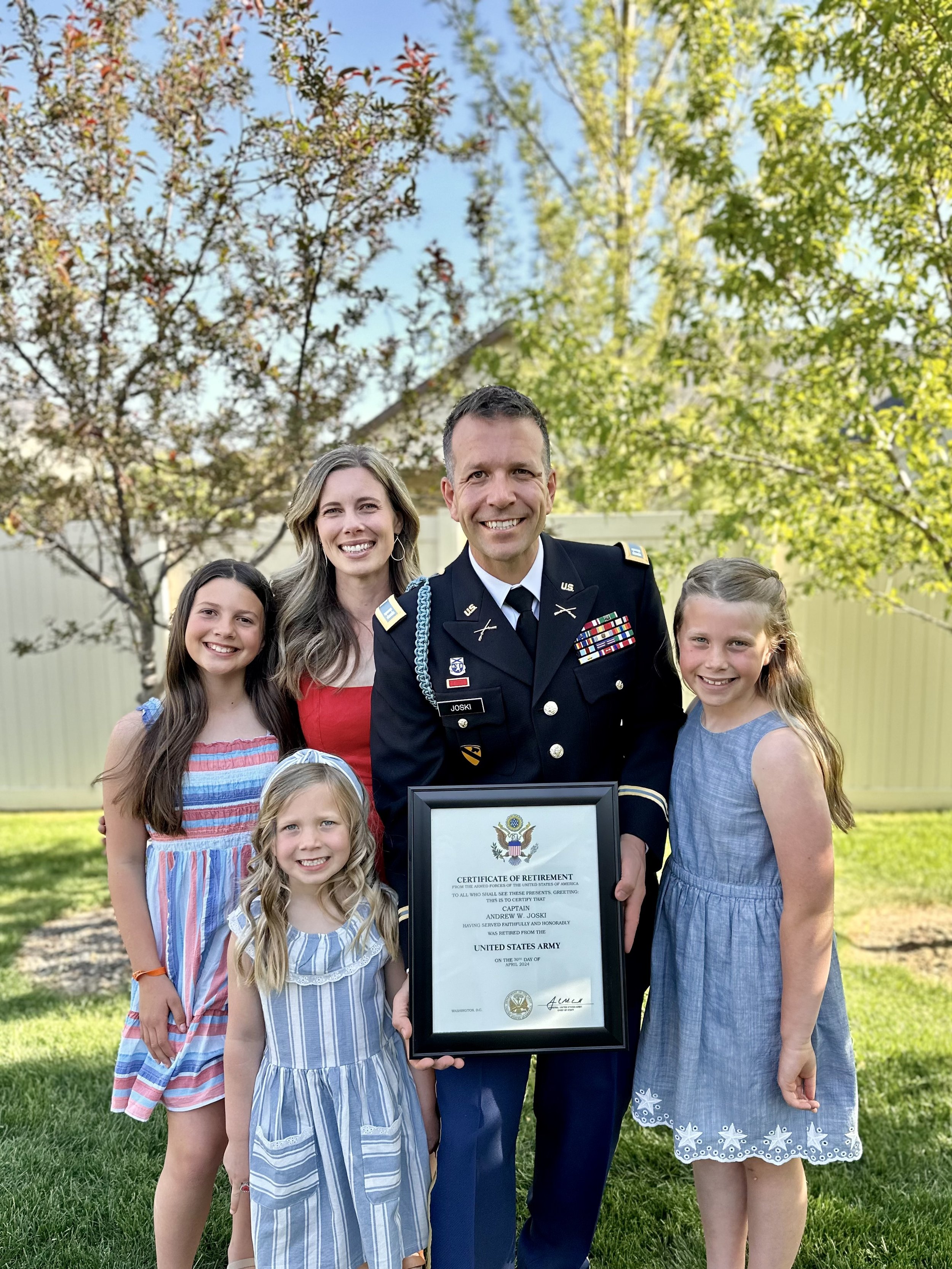 A group of six people, including a man in a military uniform, families, and children, standing outdoors in a backyard with trees and a fence, holding a framed certificate of retirement.