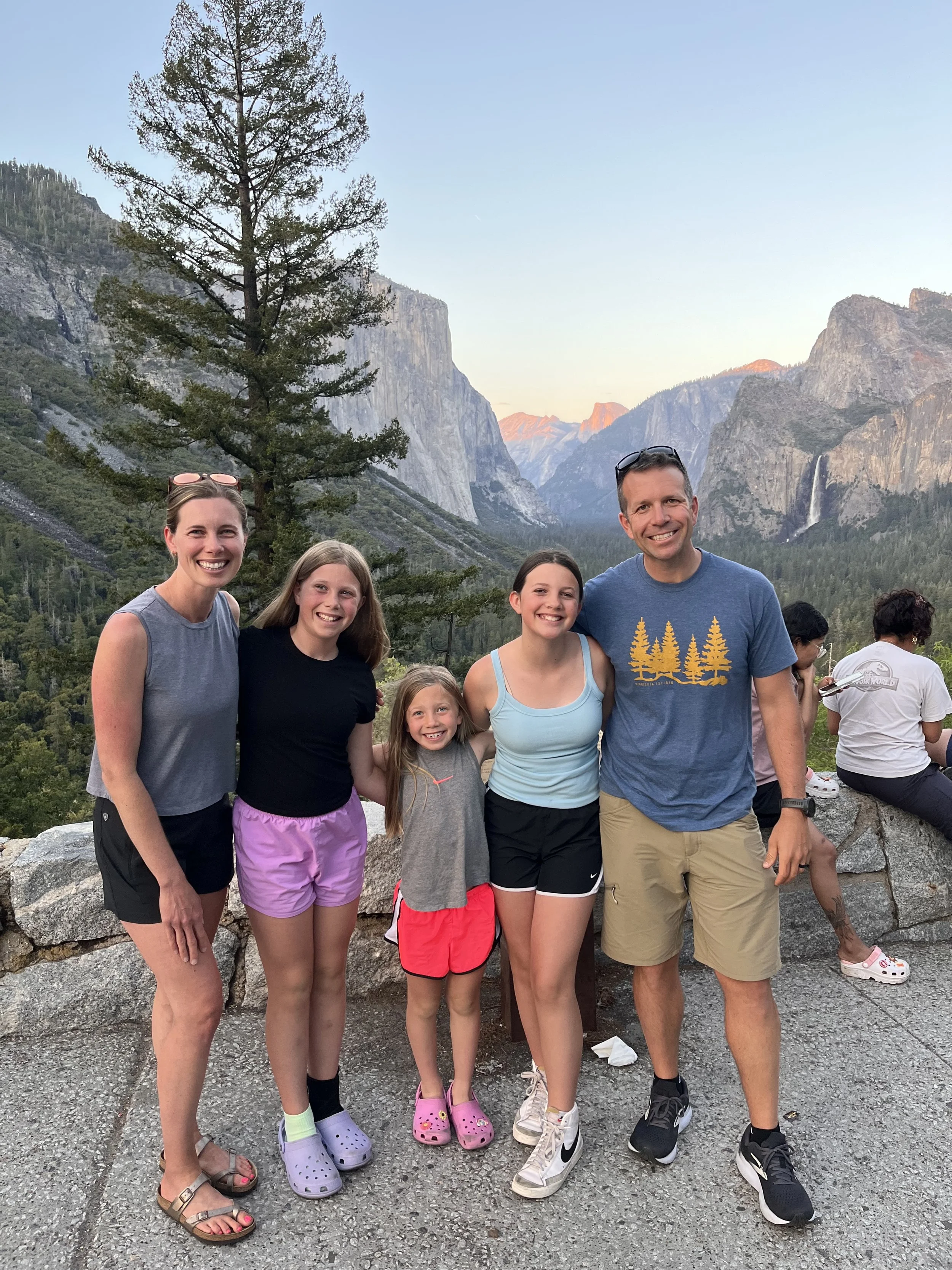 A group of five people standing on a scenic mountain overlook with tall trees and rocky peaks in the background during sunset, smiling at the camera.