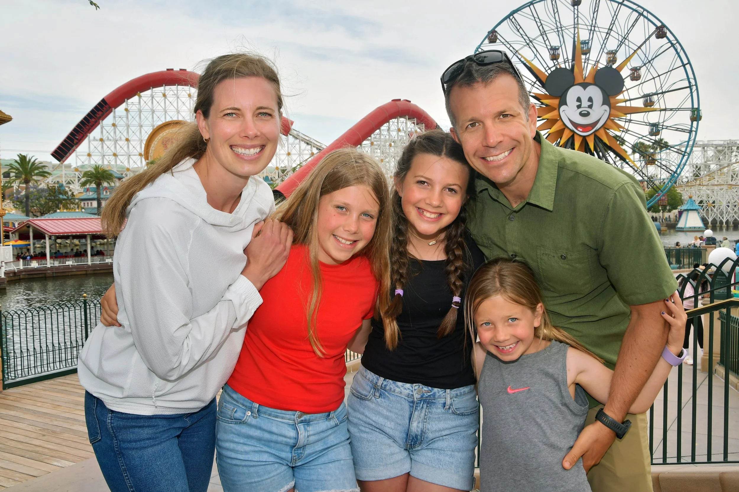 A family of five smiling and posing for a photo at an amusement park, with a roller coaster and Mickey Mouse Ferris wheel in the background.