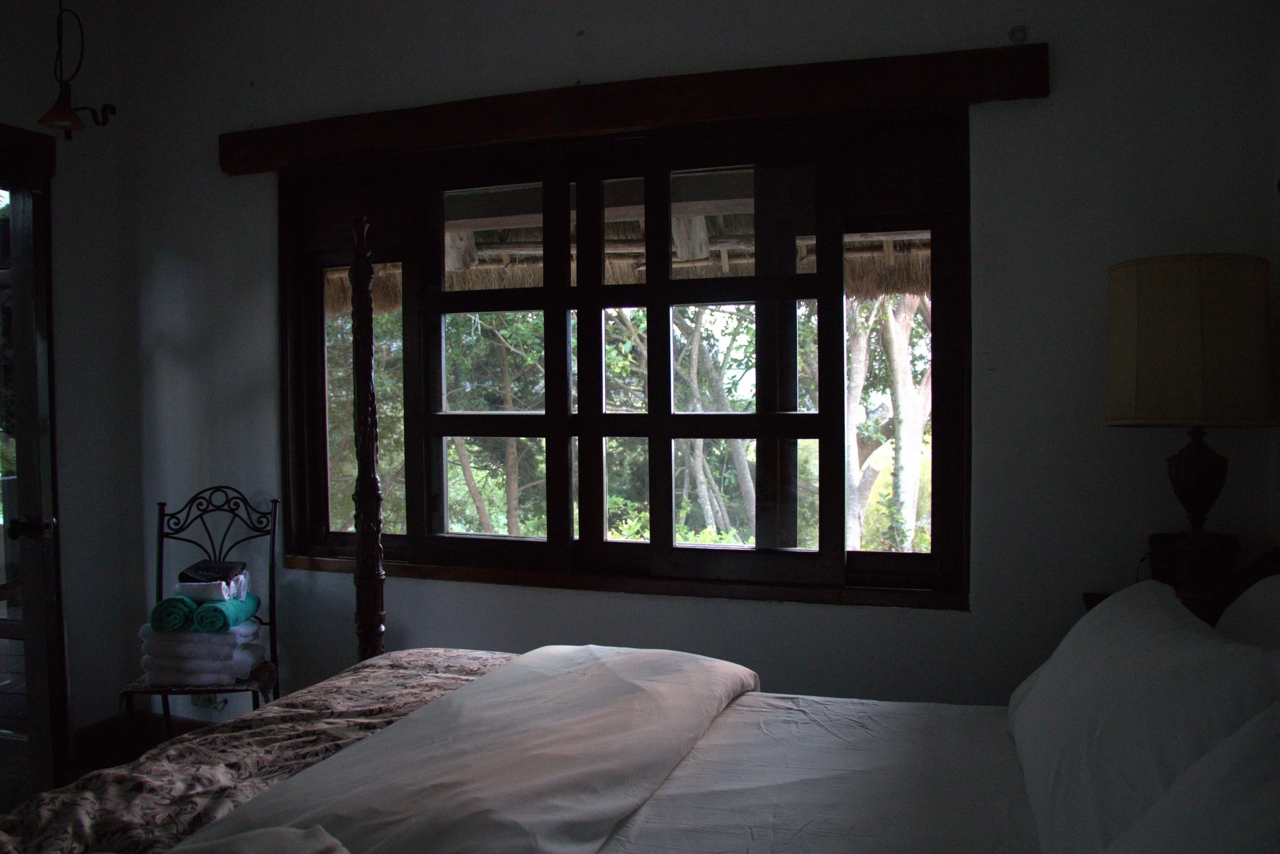 A bedroom with a bed, window view of trees, and furniture including a bedside table with rolled towels and a lamp.