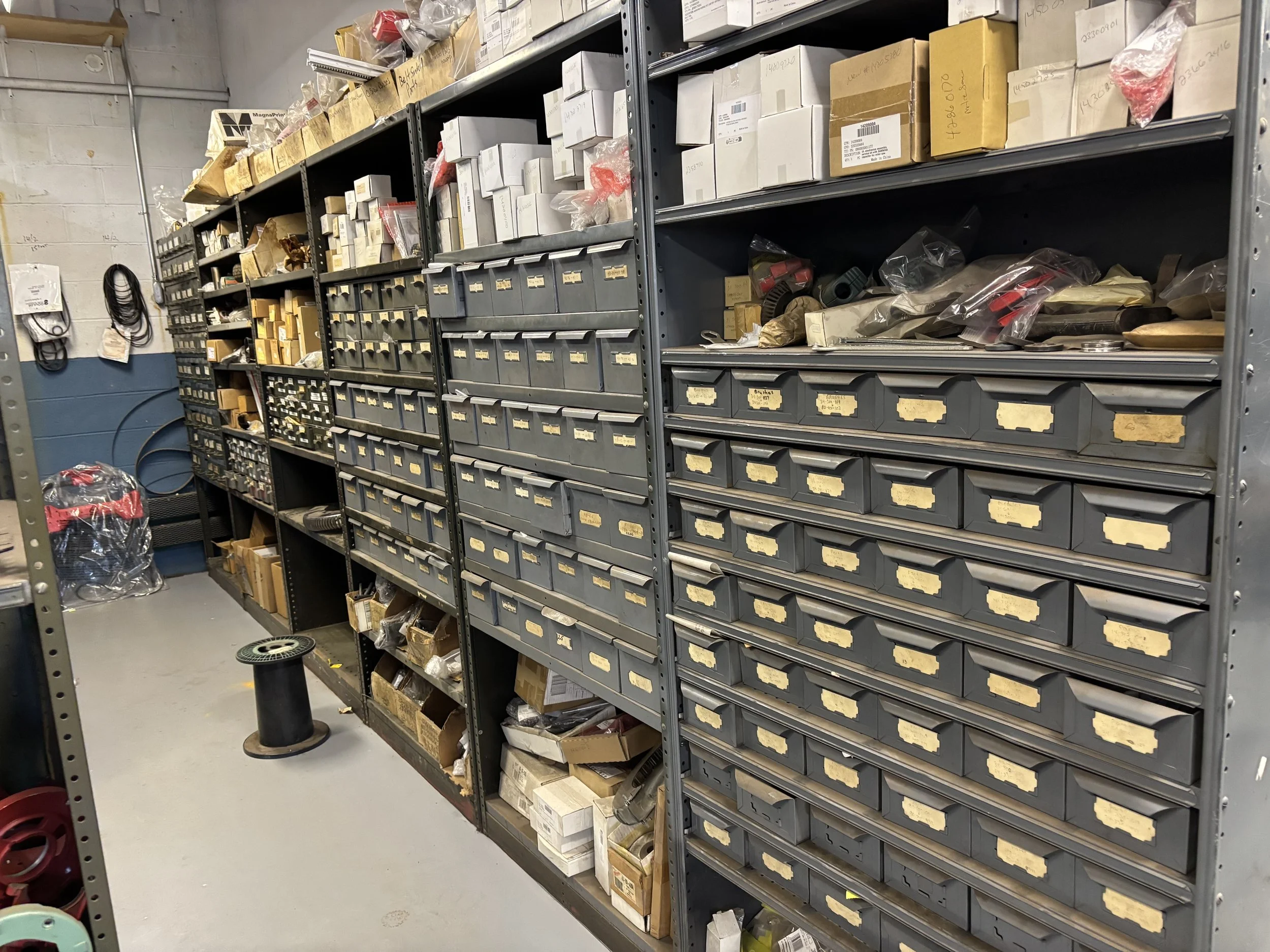 View of metal shelving units filled with boxes and plastic bags, holding various small parts and tools in a workshop or storage room.