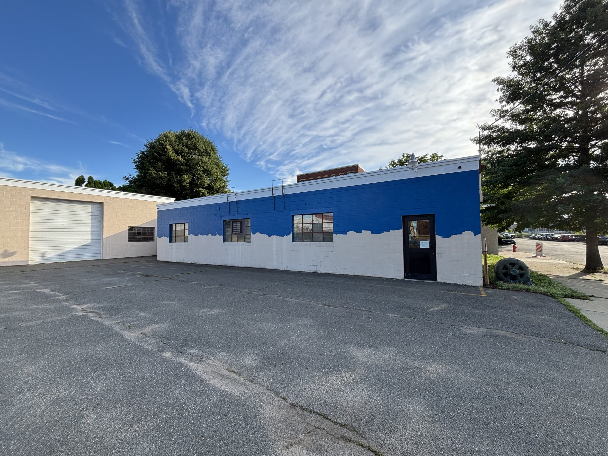 A storefront building with a blue and white color scheme, large windows, and a black door, located in a parking lot with some trees and a clear sky with clouds.