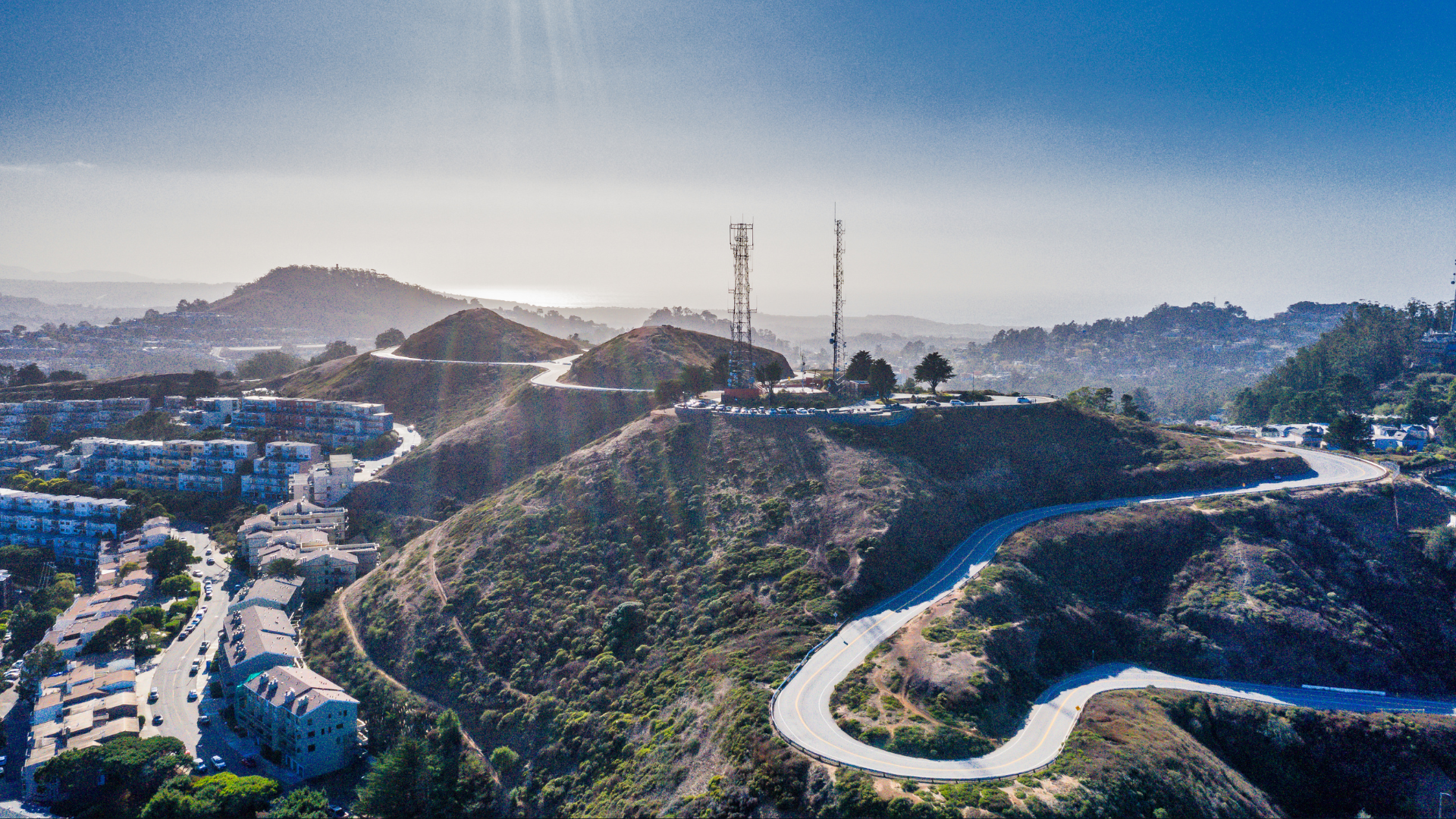 Aerial view of a hilly area with a winding road, a parking lot with numerous cars, communication towers, and a residential neighborhood at the foot of the hills.